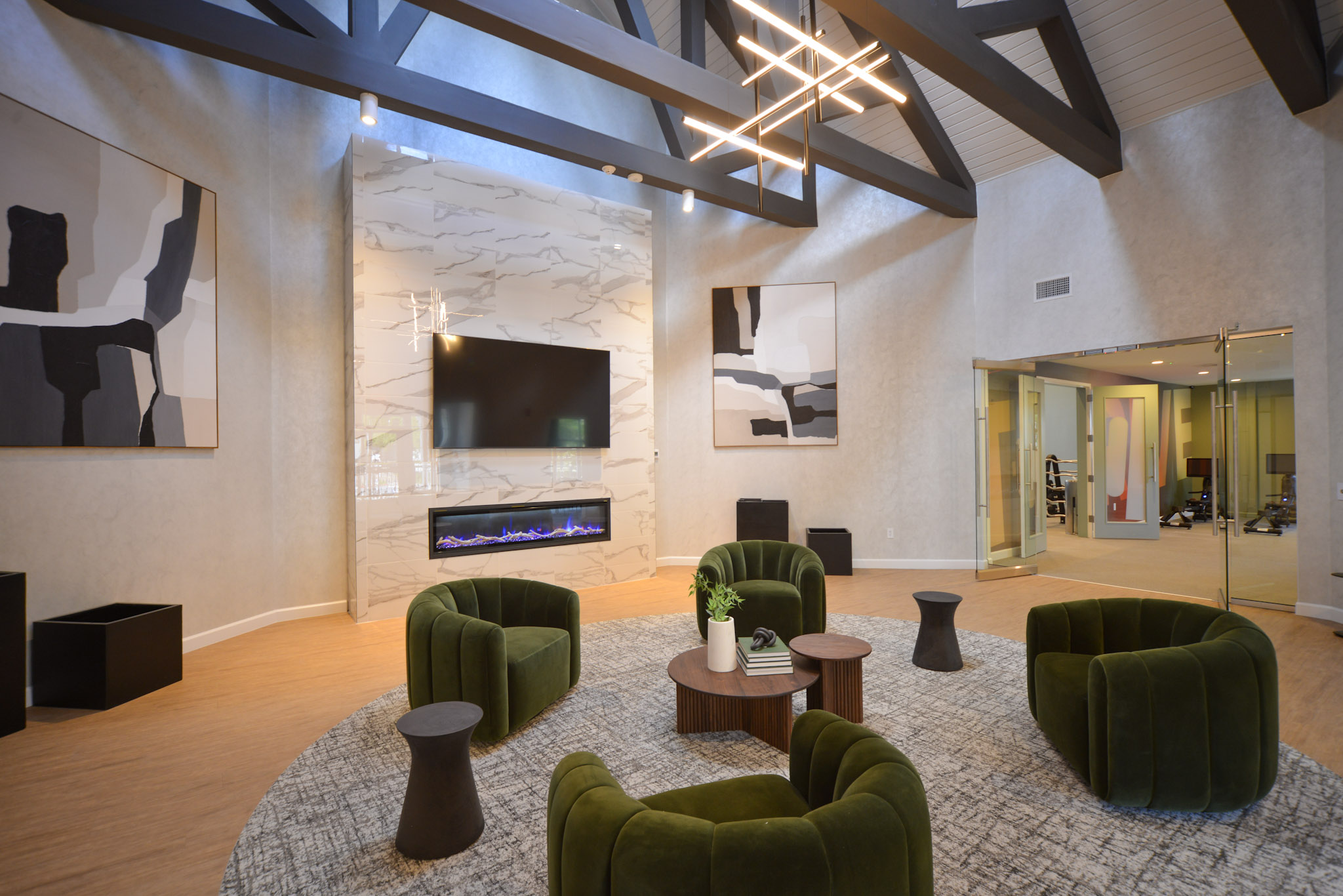 Modern living room featuring a high ceiling with dark beams, large abstract artworks on the walls, a stylish electric fireplace, and green velvet lounge chairs arranged around a wooden coffee table on a circular rug. A glass doorway leads to an adjacent room with additional furniture.