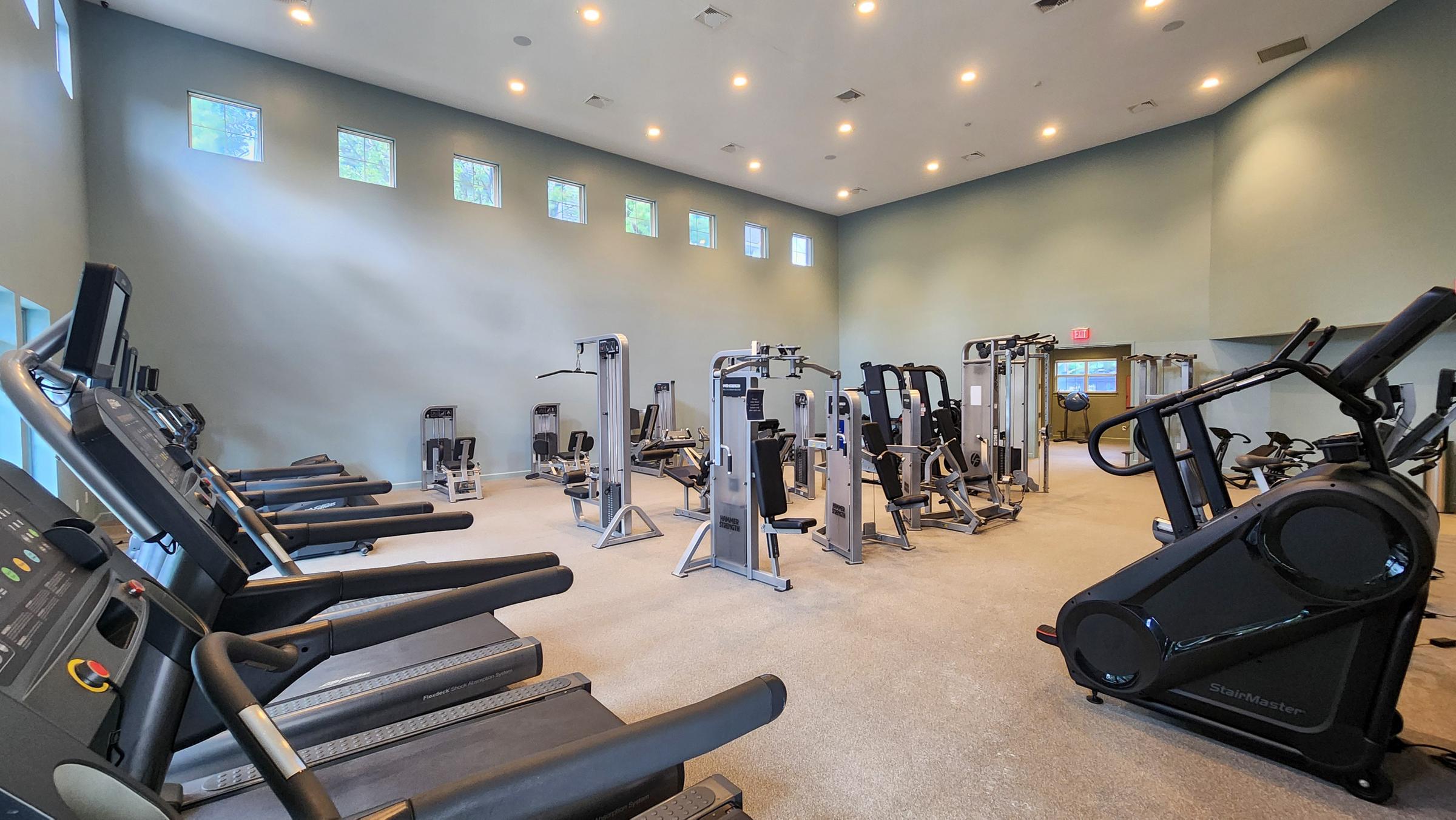 A spacious, well-lit gym featuring various exercise equipment, including treadmills, elliptical machines, and weight machines. The room has large windows allowing natural light, with a clean, modern design and a light-colored carpeted floor.
