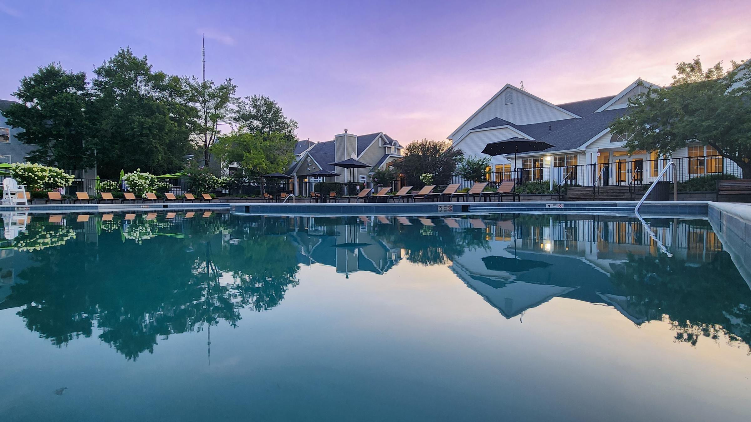 A serene swimming pool at sunset, featuring lounge chairs around the water. The sky is a gradient of purple and orange, reflecting on the calm surface of the pool. In the background, a well-maintained building with multiple windows is visible, surrounded by trees and landscaping.