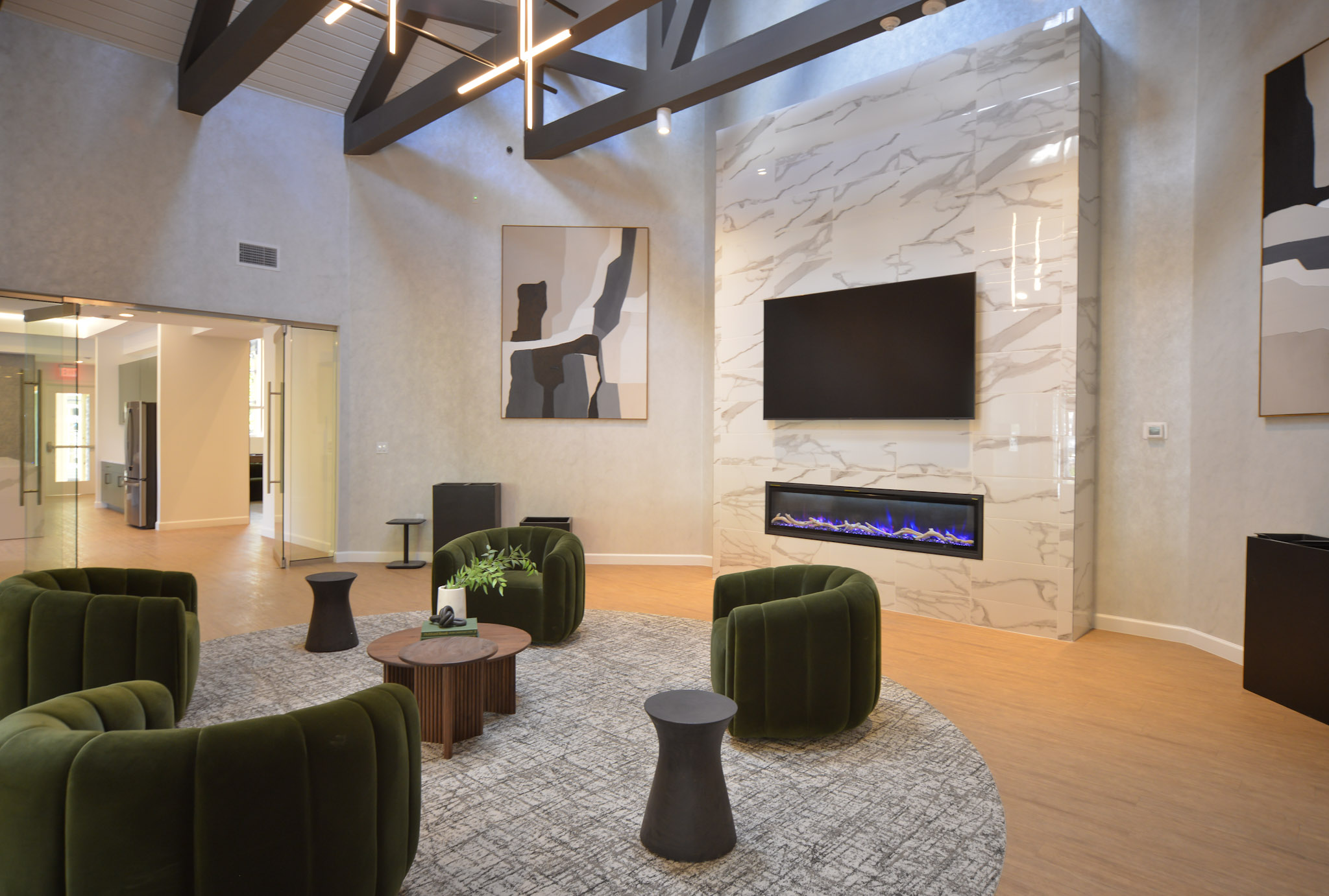 A modern lounge area featuring stylish green velvet chairs arranged around a circular rug. A sleek coffee table sits in the center, with a decorative plant. To one side, a contemporary marble fireplace with an electric insert and a large flat-screen TV hang on the wall. Abstract artwork decorates the walls.
