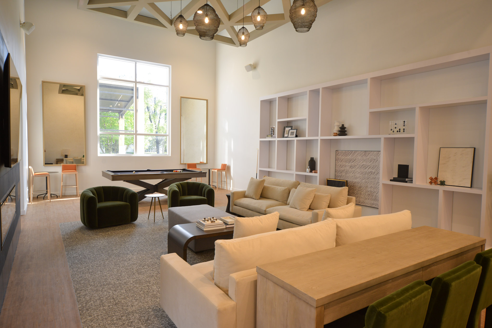 A modern living room featuring a cozy arrangement of green and beige sofas, a wooden coffee table, and a light-colored dining table. The space is illuminated by pendant lights and has large windows allowing natural light in. There are decorative shelves and a pool table in the background, enhancing the stylish ambiance.
