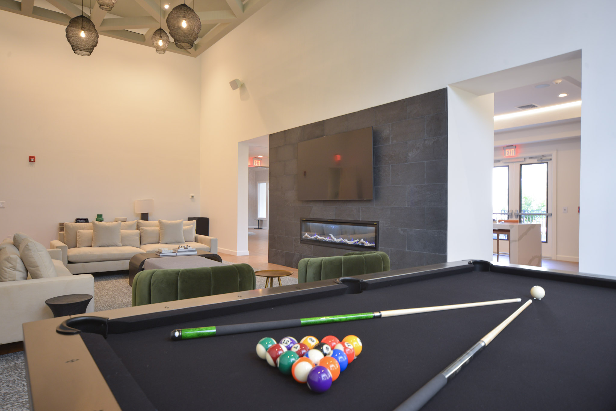 A modern interior featuring a pool table with balls arranged in a triangle, cues resting on the table. In the background, a cozy seating area with a light-colored sofa and a wall-mounted television above a sleek fireplace. Large windows provide natural light, enhancing the spacious vibe.