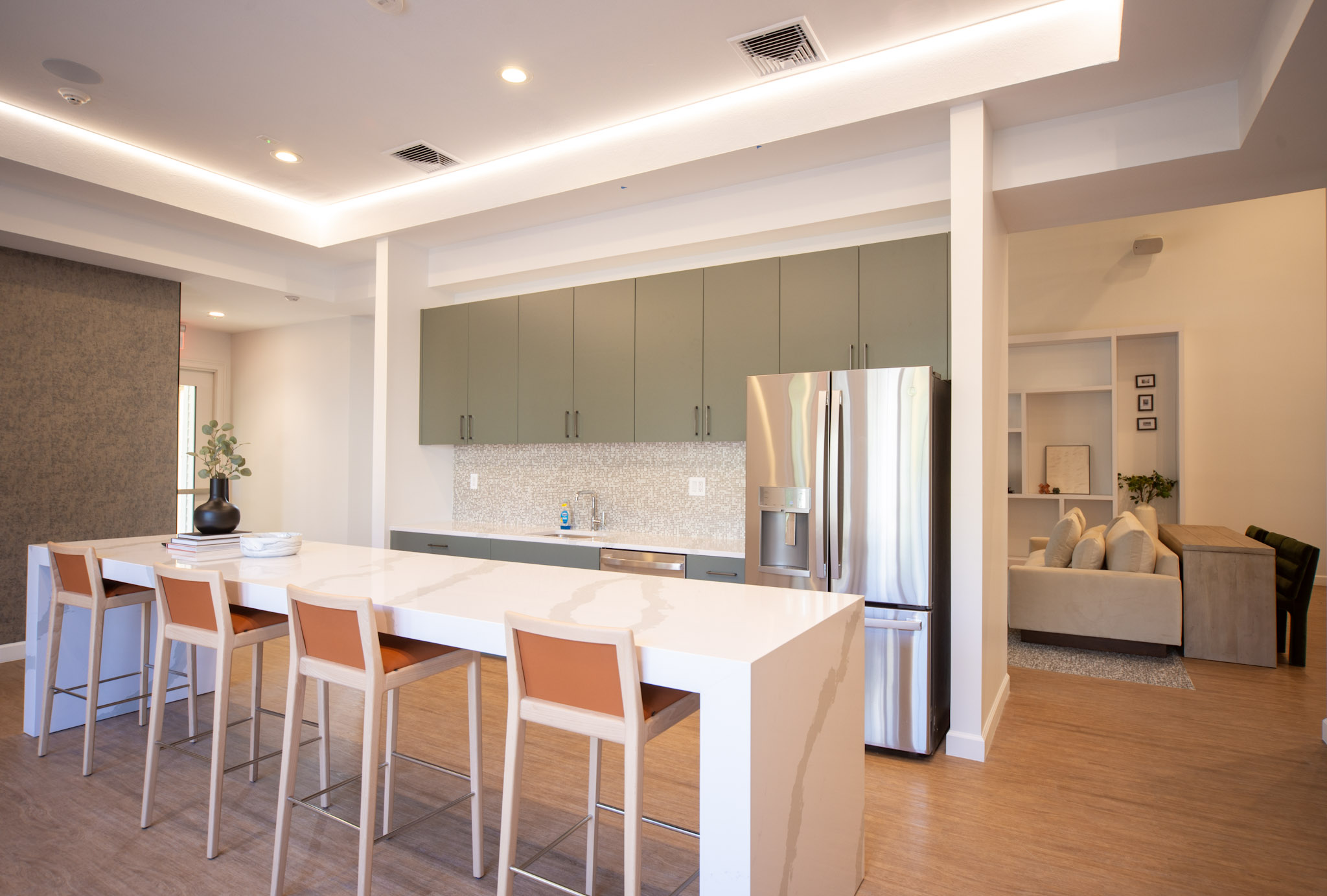Modern kitchen featuring a large white countertop island with orange bar stools, stainless steel refrigerator, and green cabinetry. The space is well-lit with recessed lighting and has a cozy living area visible in the background, showcasing a comfortable sofa and decorative elements.