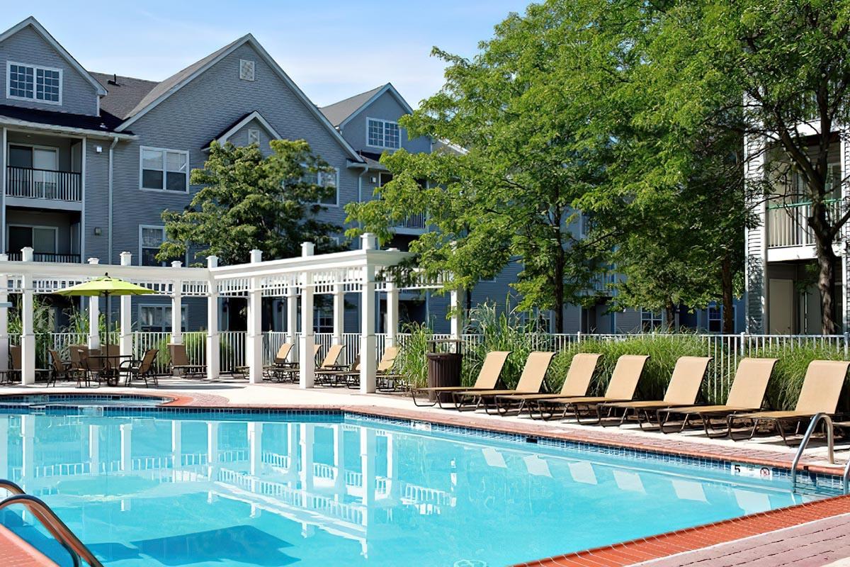 Outdoor swimming pool surrounded by lounge chairs and umbrellas, with a landscaped area of trees and shrubs. In the background, there are residential buildings with balconies. The scene is bright and inviting, perfect for relaxation on a sunny day.