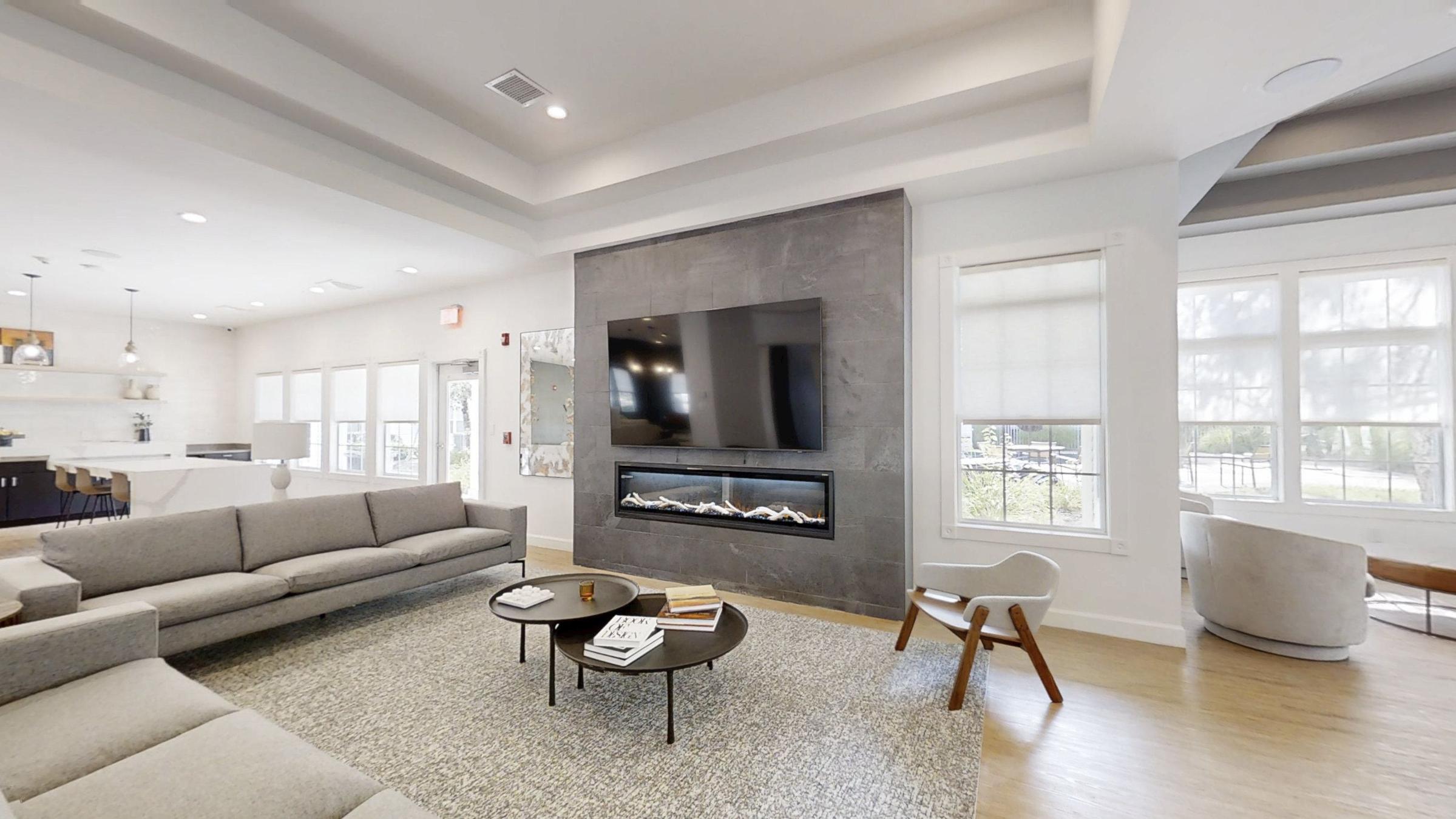 Modern living room featuring a gray sectional sofa, circular coffee table with magazines, and a stylish fireplace unit beneath a large flat-screen TV. Bright windows allow natural light to fill the space, complemented by a neutral color palette and contemporary decor.