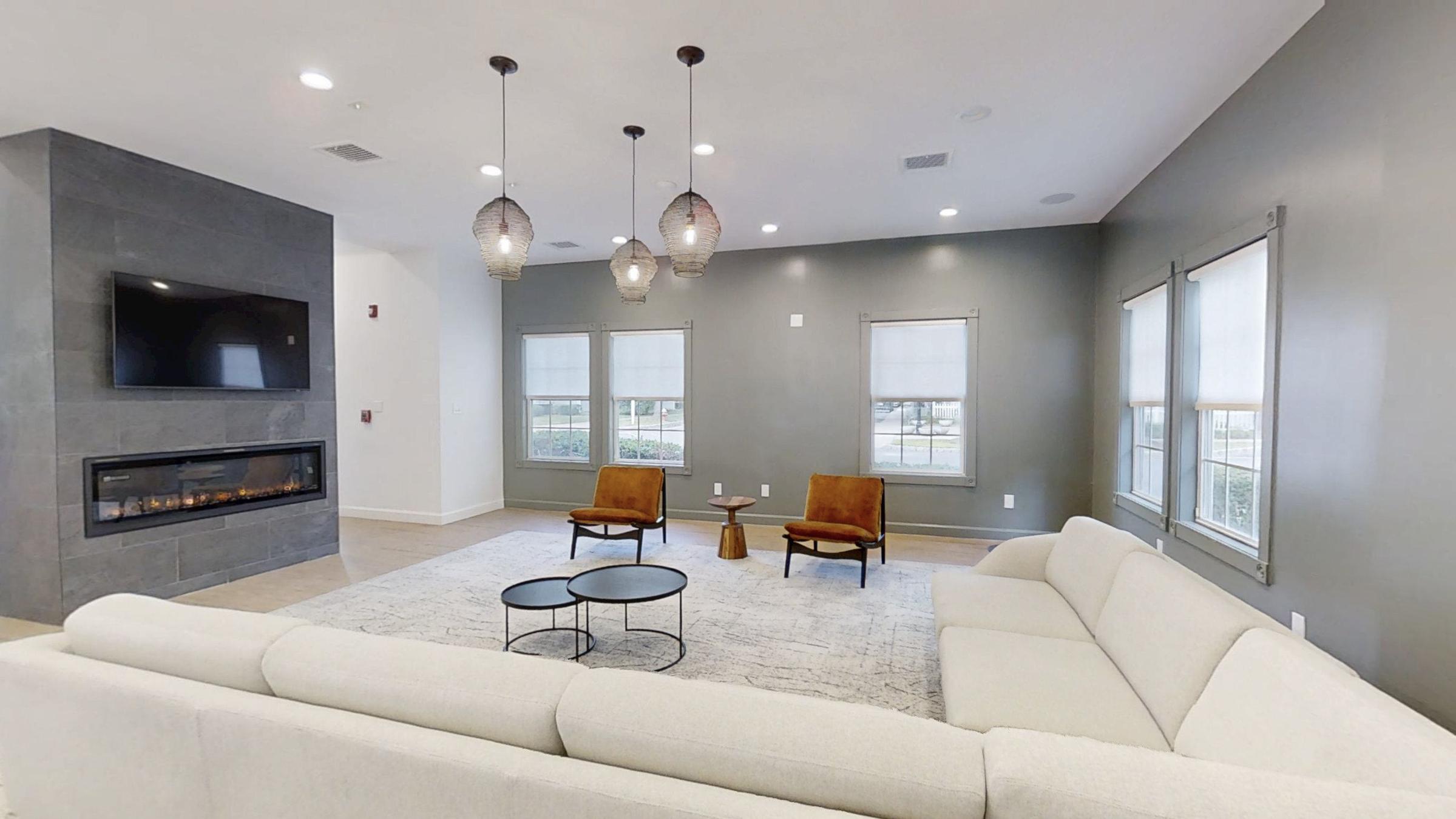 A modern living room featuring a light beige sectional sofa, two orange accent chairs, and a round coffee table. There’s a wall-mounted TV above a contemporary fireplace, with decorative pendant lights hanging from the ceiling. Large windows provide natural light, enhancing the space's inviting atmosphere.
