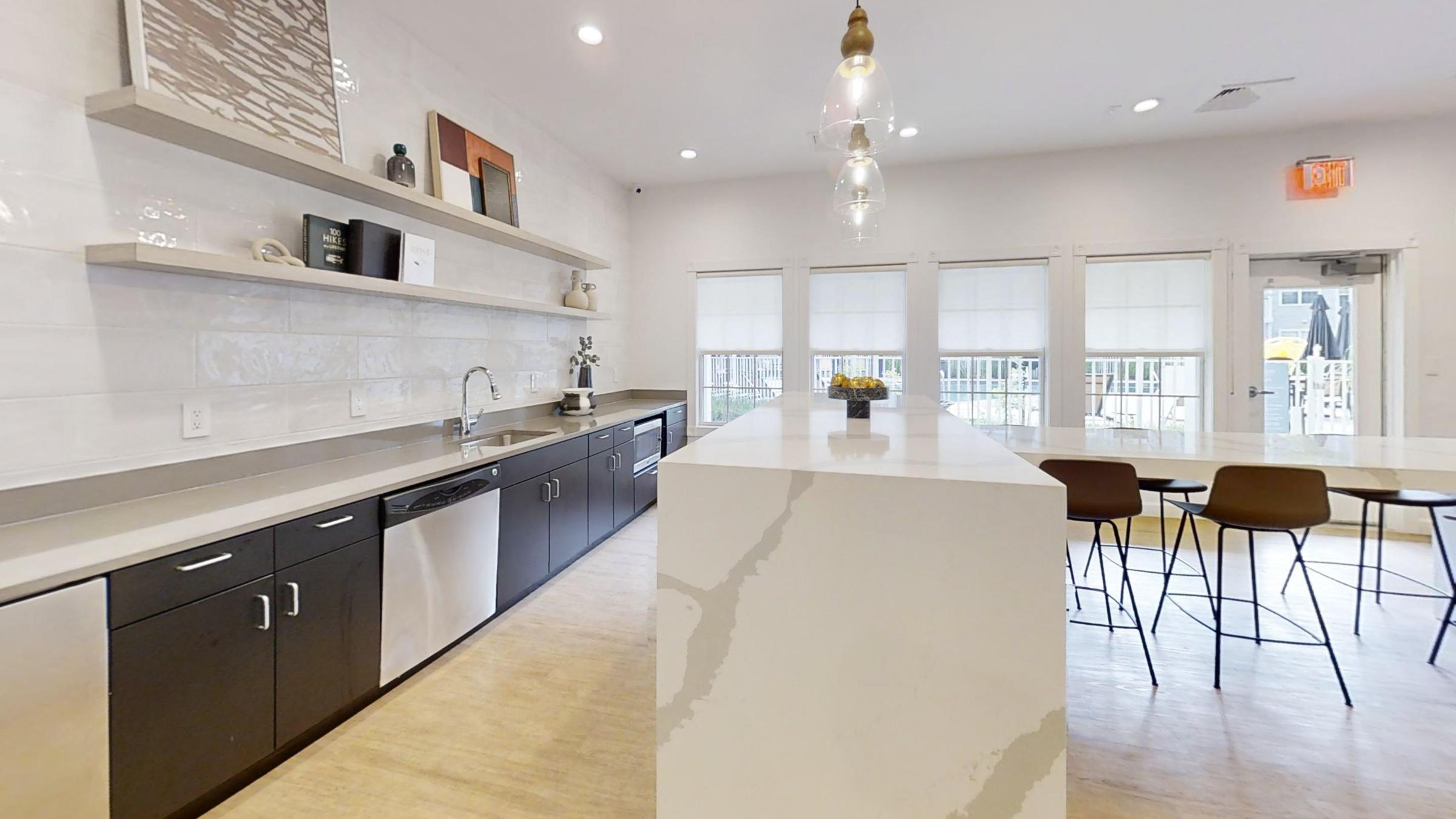 Modern kitchen with a large white island, dark cabinetry, and stainless steel appliances. The walls are adorned with minimalist decor, and there’s a dining area with several brown chairs. Natural light floods the space through large windows, creating an inviting atmosphere.