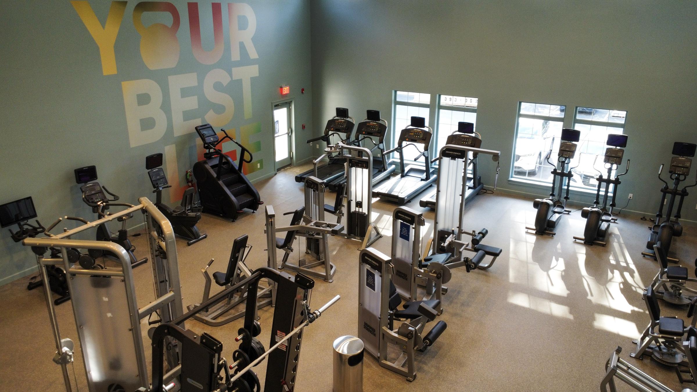 A spacious gym interior featuring various exercise machines, including treadmills, ellipticals, and strength training equipment. The walls are painted in calming colors, with the phrase "YOUR BEST LIFE" prominently displayed in bold letters. Large windows allow natural light to fill the space.