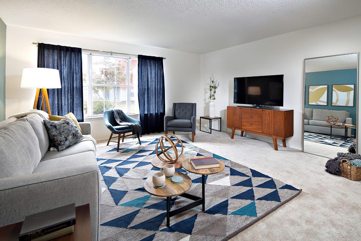 A modern living room featuring a light gray sofa with decorative pillows, a round coffee table with books and cups, a patterned area rug, and a dark blue armchair. Large windows with navy curtains let in natural light, and a TV mounted on the wall is facing a mid-century modern wooden TV stand. A mirror reflects the room, enhancing the light and space.