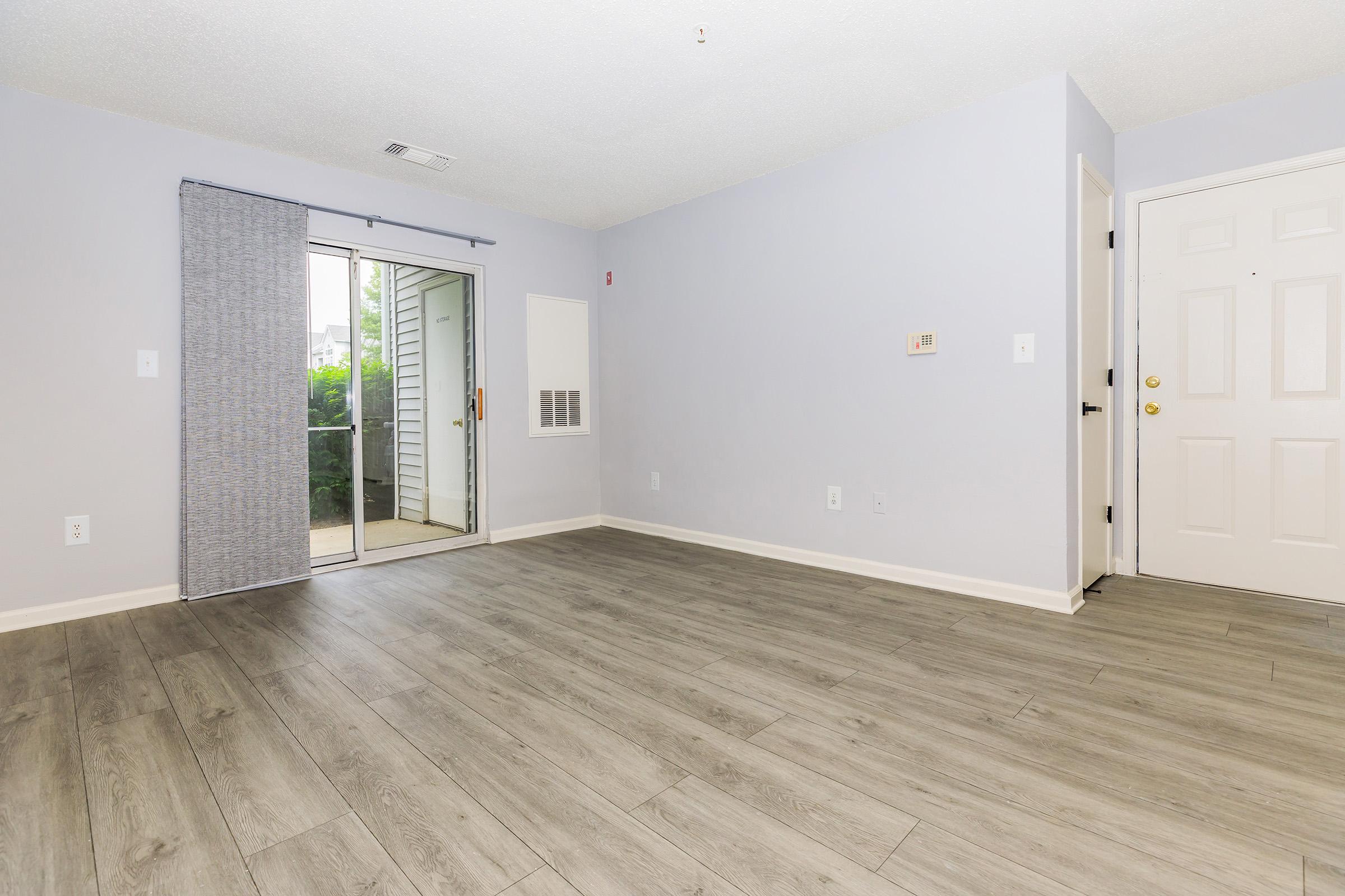 Empty room with light gray walls and vinyl flooring. A sliding glass door leads to a small balcony, and there’s a door on the right. The space is well-lit, featuring a simple, clean aesthetic with no furniture. It has a modern, minimalist feel, suitable for various decorating styles.