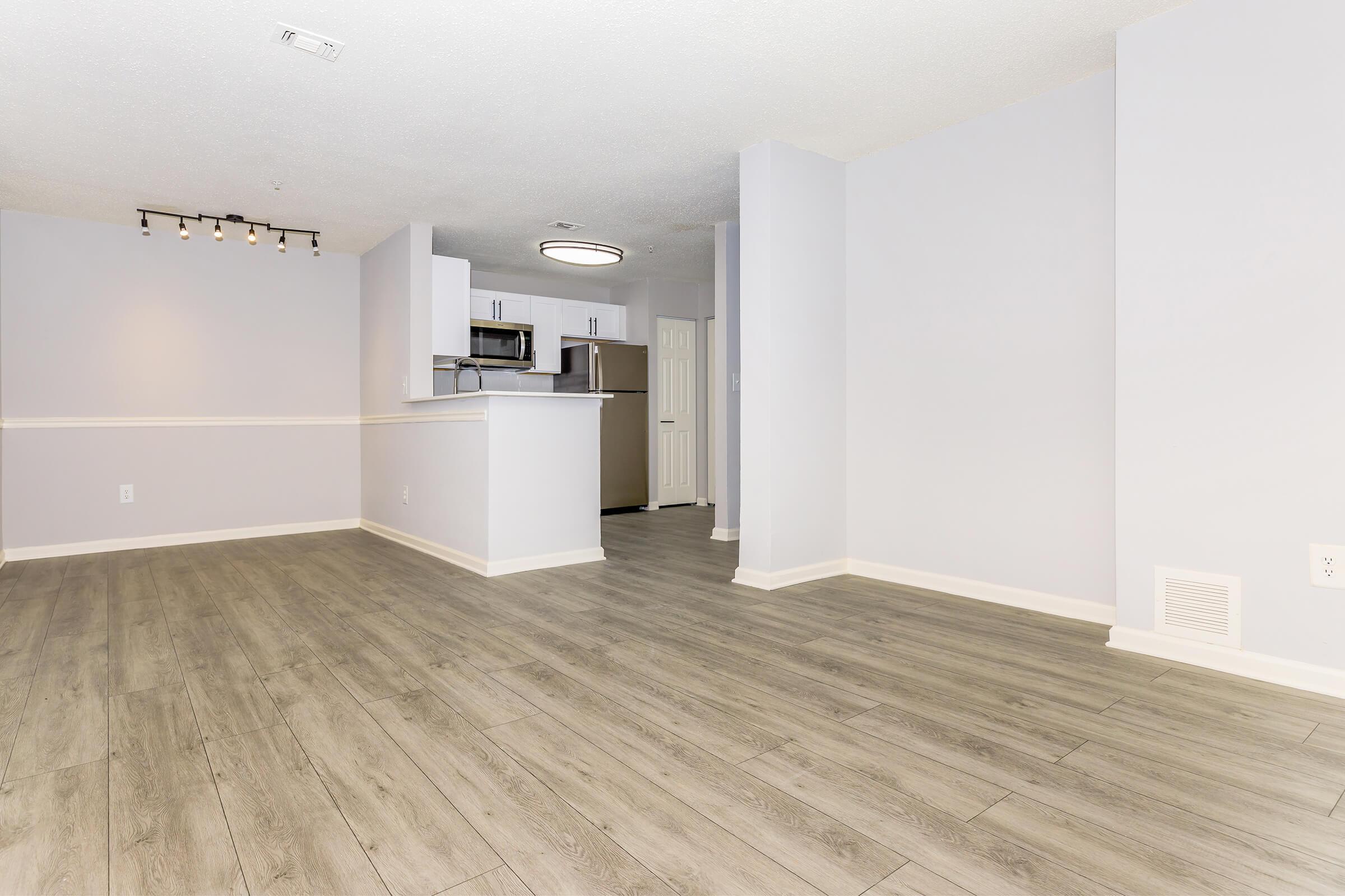 A spacious, vacant living area featuring light gray walls and new, light wood flooring. The room has an open layout that connects to a modern kitchen with stainless steel appliances. Natural light fills the space, and there is recessed lighting on the ceiling.