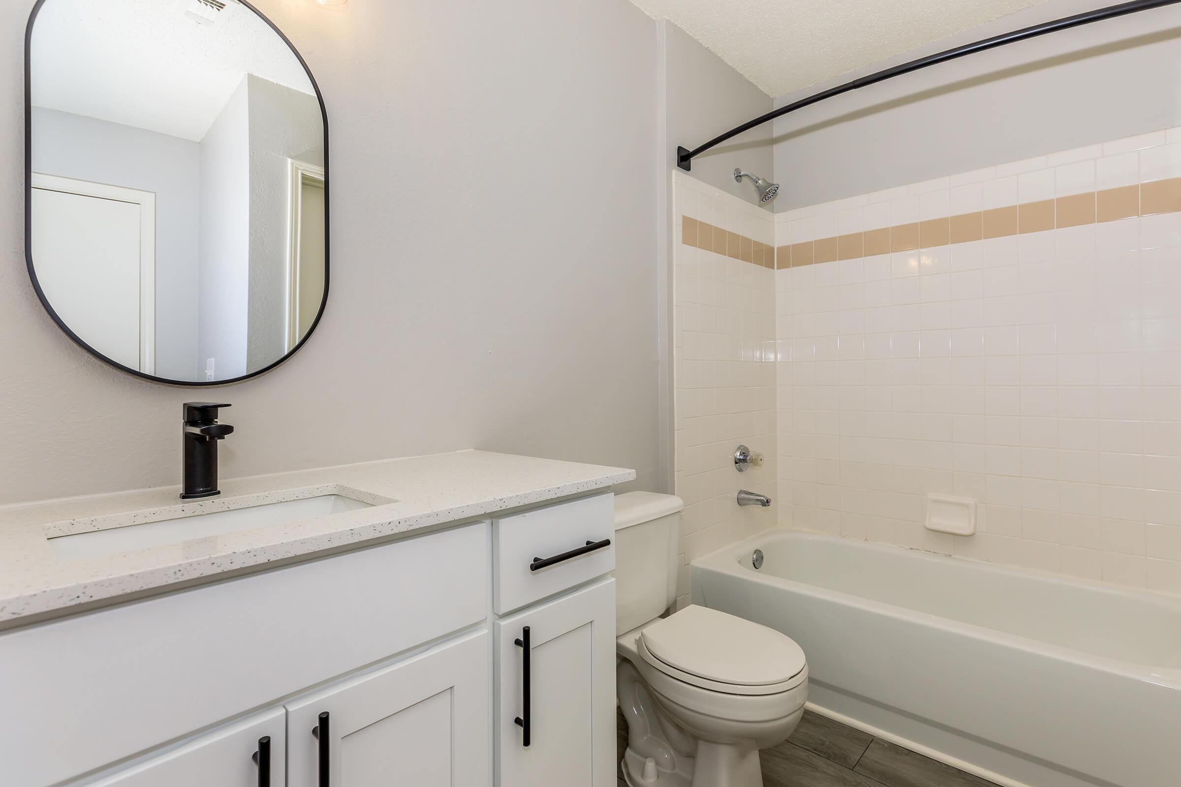 A modern bathroom featuring a white bathtub with a shower, a round mirror above a white sink with a minimalist faucet, and cabinetry with black handles. The walls are painted light gray, and the tiled shower area includes a subtle beige accent. Natural light enhances the bright, clean aesthetic.