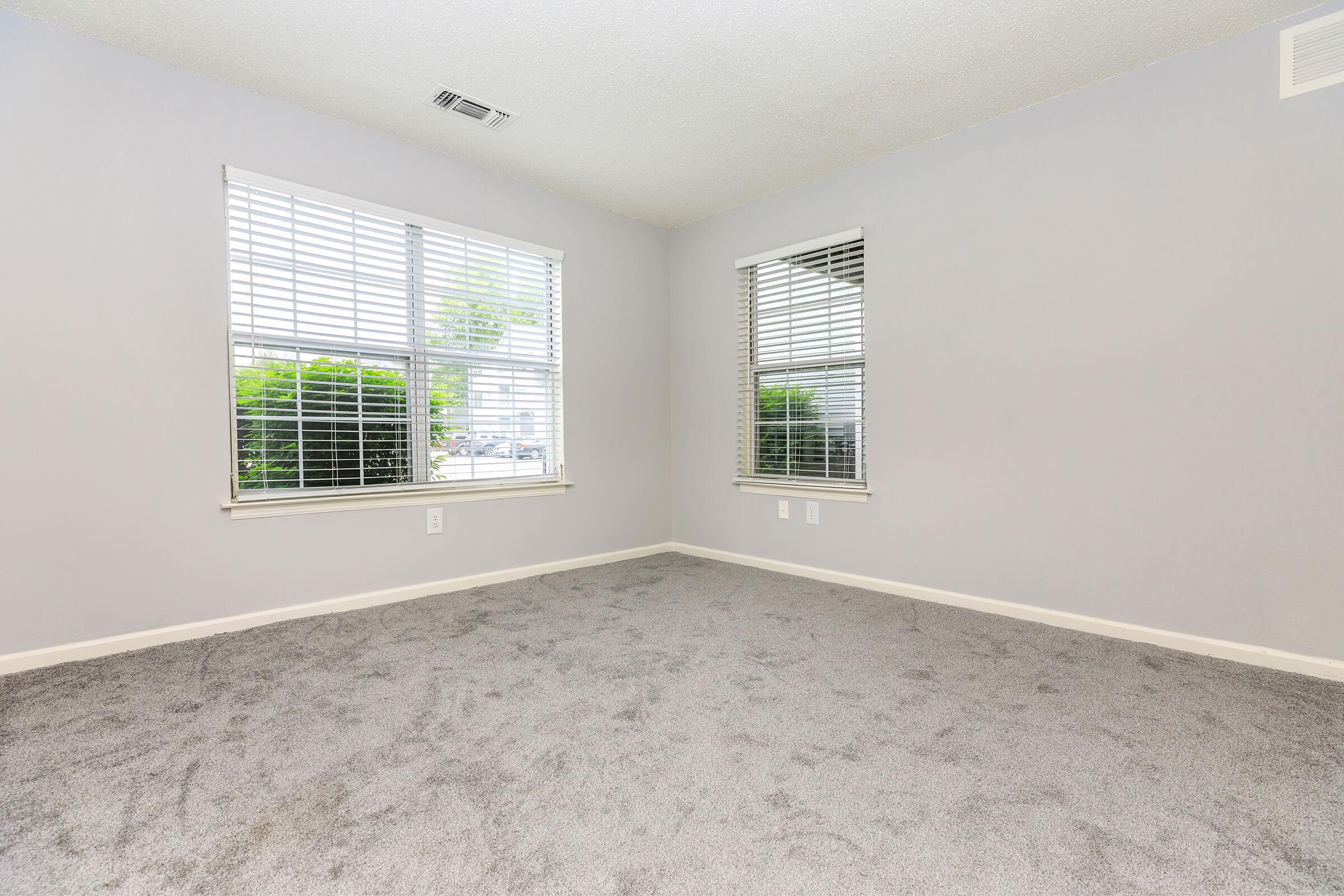 A vacant room with light gray walls and a plush gray carpet. Two windows feature white blinds and allow natural light, revealing greenery outside. The space is empty, creating a clean and neutral atmosphere, suitable for various uses or decoration.