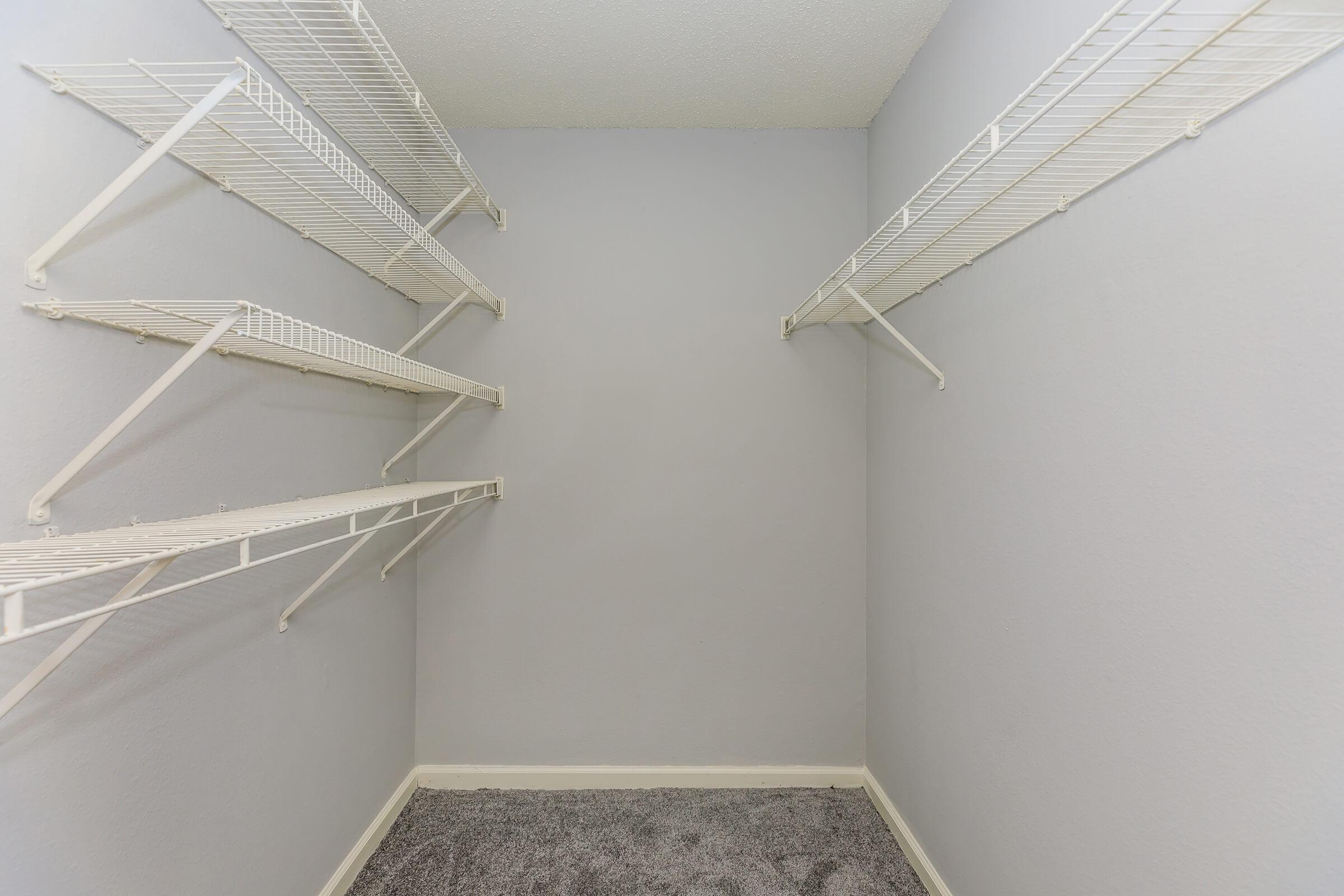 Empty closet with gray walls and carpeted floor, featuring white wire shelving mounted on the walls. The space is clear of any items, providing a blank canvas for storage or organization.