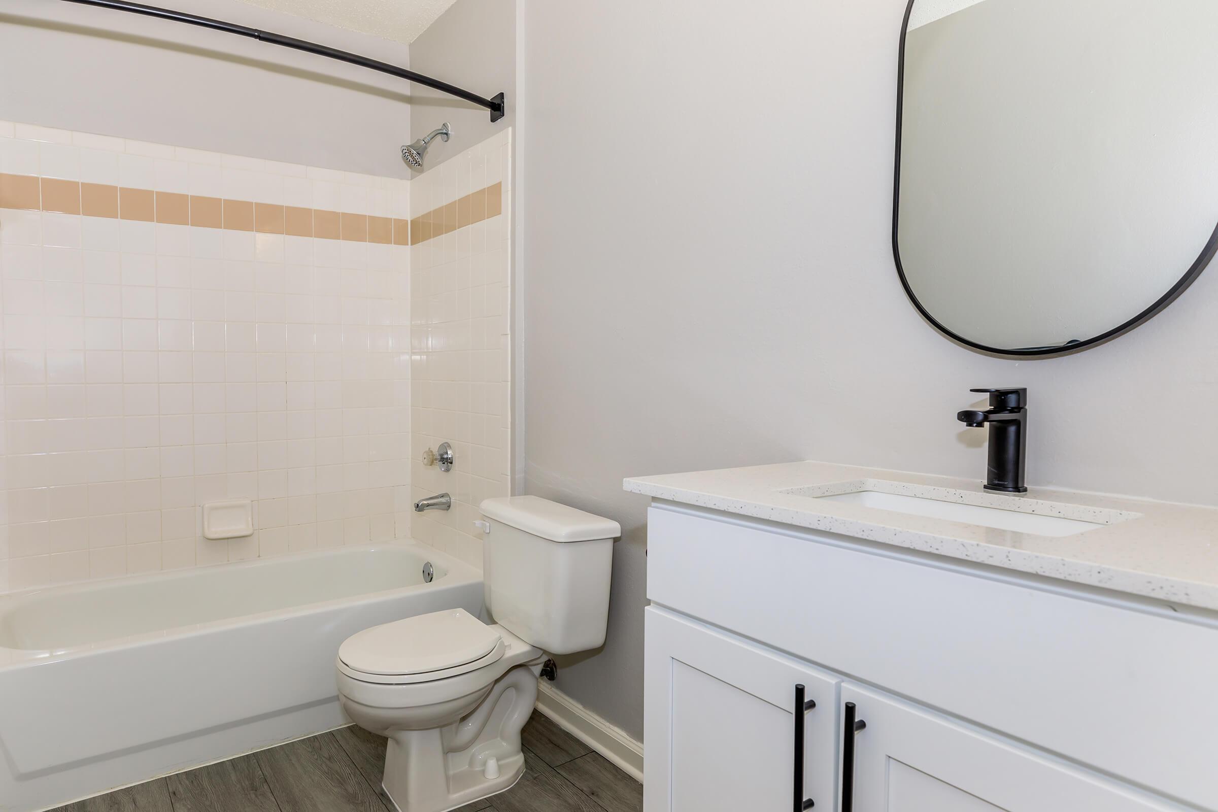 A modern bathroom featuring a white bathtub with a showerhead, a toilet, and a sleek vanity with a black faucet. The walls are light gray, and there's a large, round mirror above the sink. The tiled backsplash has a pale orange accent strip, contributing to a clean, minimalist aesthetic.