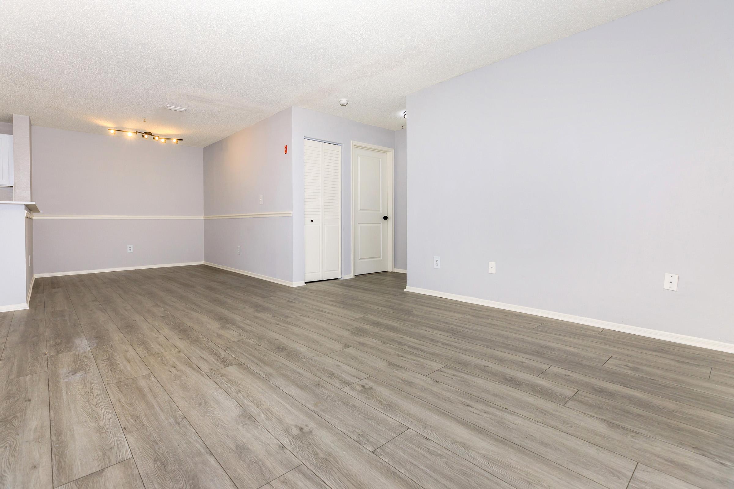 A spacious, well-lit room with light gray walls and light wood flooring. The area is empty, featuring a doorway with a closed white door on one side and a partially visible kitchen area in the background. The ceiling has recessed lighting, creating a modern, open atmosphere.