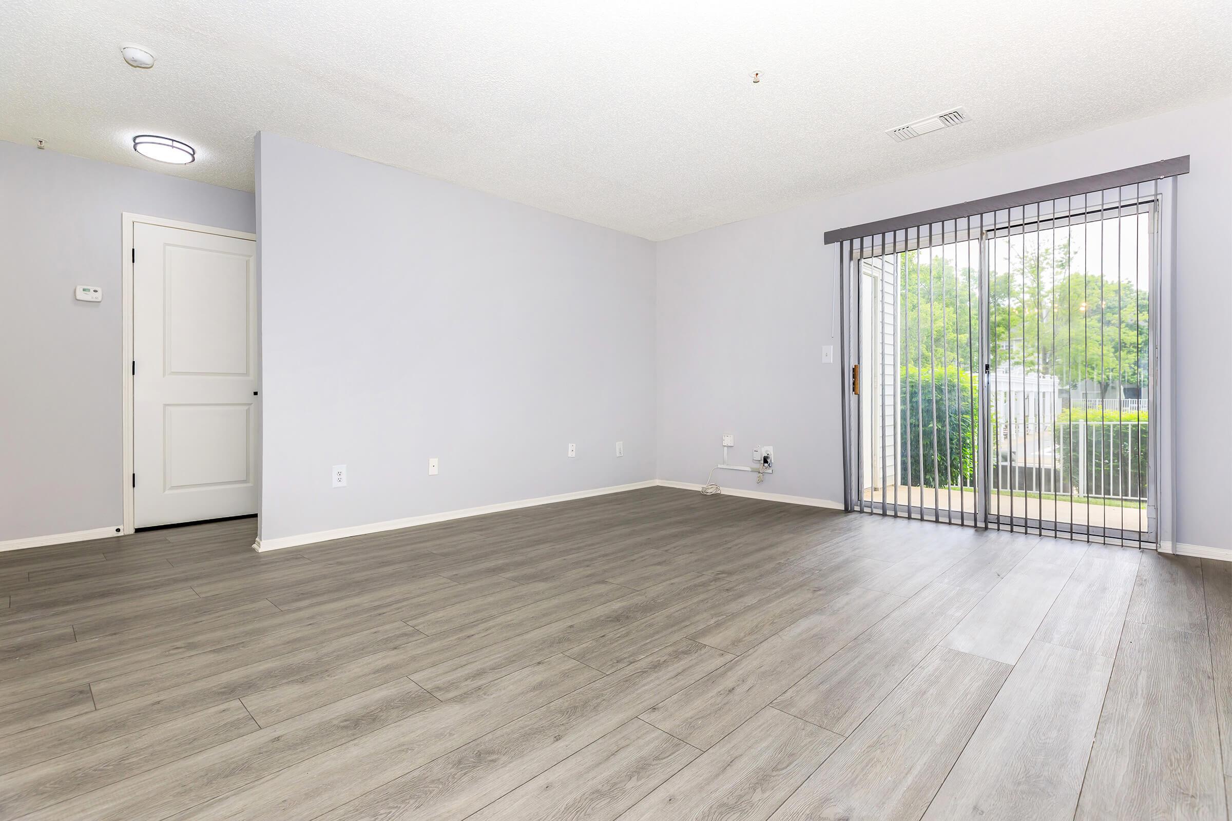 An empty room with light gray walls and wood-like flooring. There is a sliding glass door with vertical blinds leading to an outdoor area, and a nearby white door. The space is well-lit with natural light coming through the door.