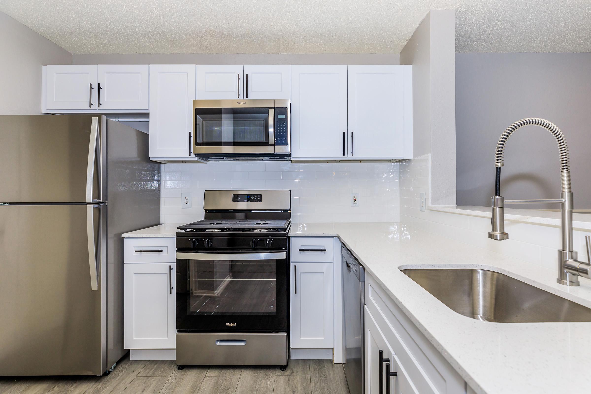 Modern kitchen featuring white cabinetry, stainless steel appliances including a refrigerator, oven, and microwave, a double sink, and a neutral-tone backsplash. The space is well-lit with natural light and has a sleek, contemporary design.