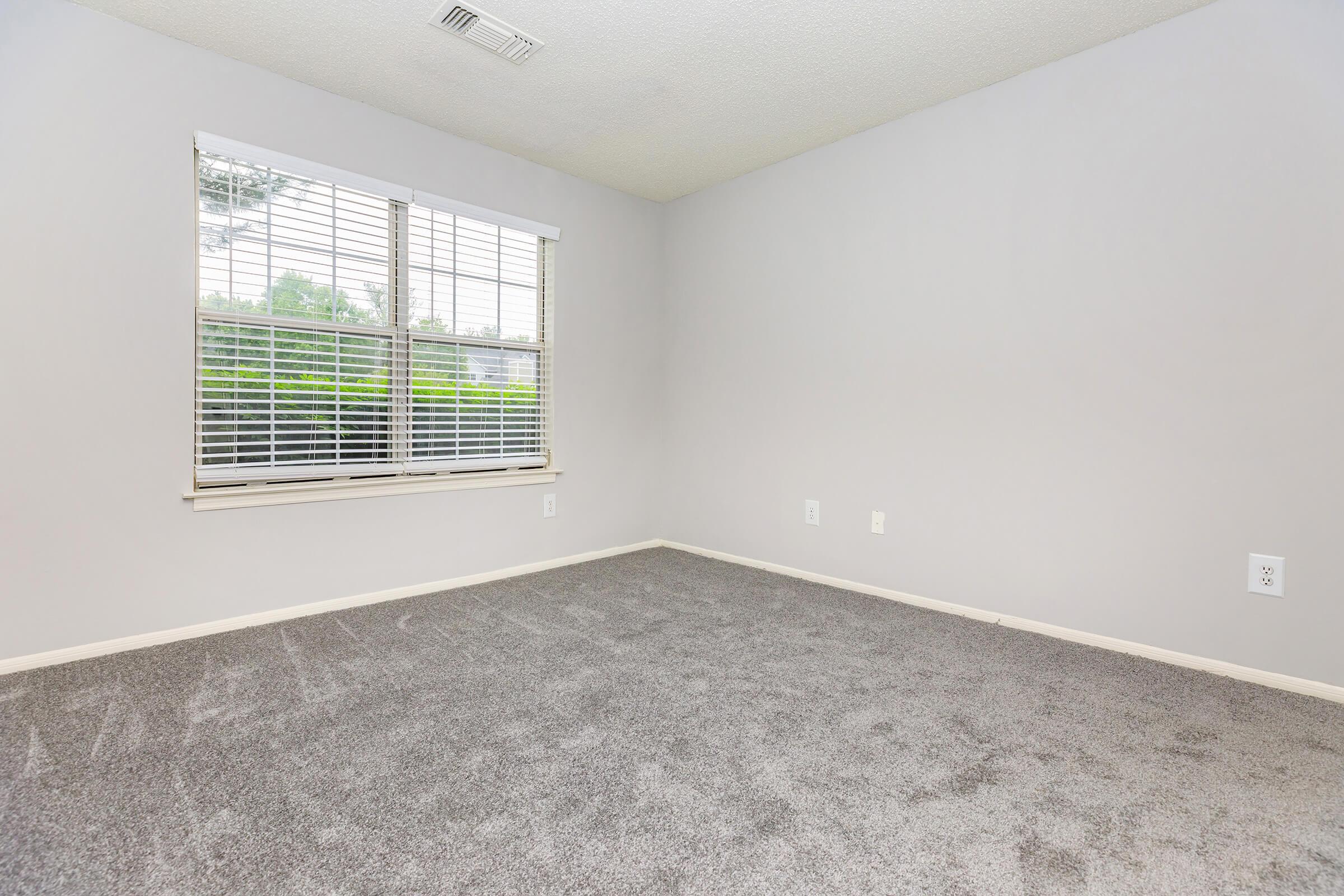 A spacious, empty room featuring light gray walls and a large window with white blinds. The floor is covered in gray carpet, and there are no furnishings or decorations. The room has a clean, minimalist appearance with ample natural light coming through the window.