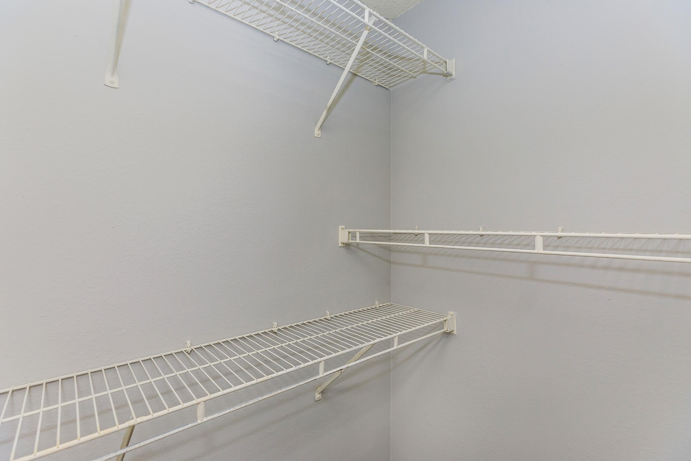 Empty closet space featuring white wire shelving along the walls. The walls are painted a light gray, creating a neutral backdrop. There are multiple shelves installed at varying heights for storage, but no items are placed on the shelves. The overall appearance is clean and organized.