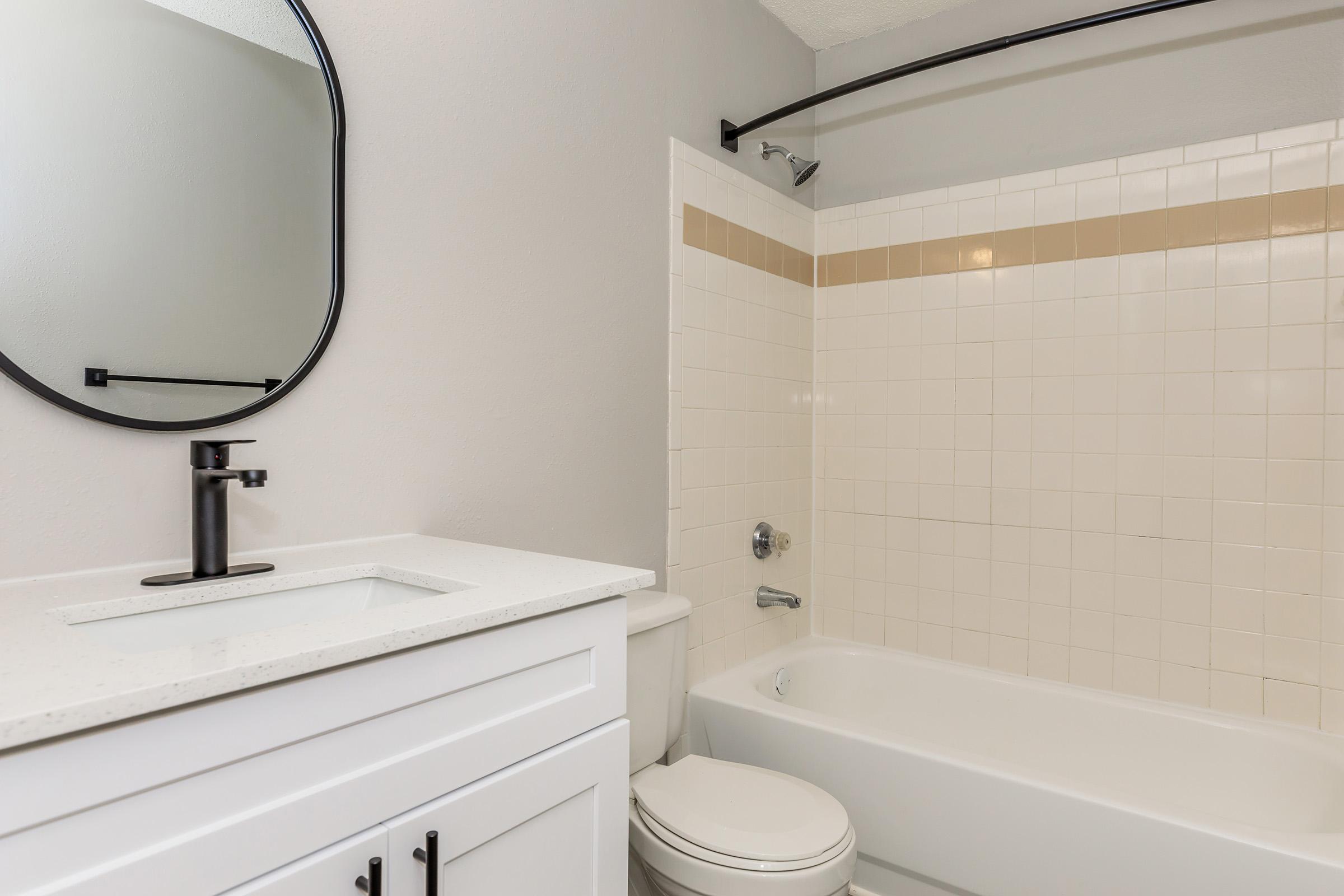 A modern bathroom featuring a white countertop sink with a black faucet, a large round mirror above it, a bathtub with a showerhead, and neutral tiled walls. The space is well-lit with a clean and minimalist design, showing a simple toilet beside the bathtub.