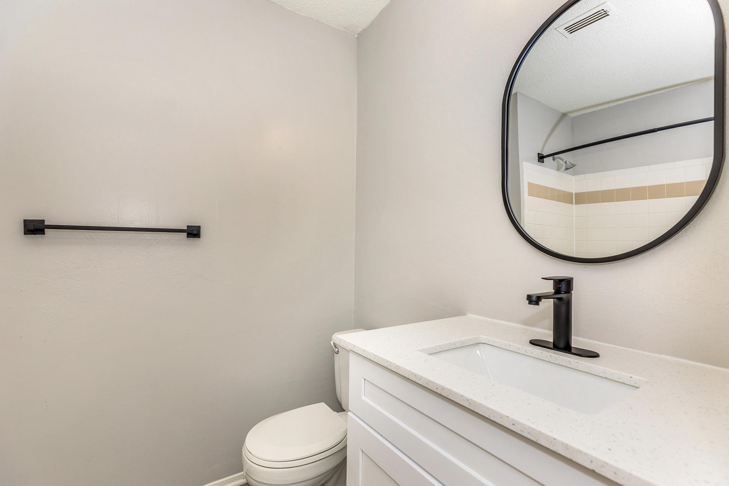 A modern bathroom featuring a round mirror above a white sink with a black faucet. To the left, there is a toilet, and a black towel bar on the wall. The walls are painted in light gray, and the shower area is partially visible with neutral tiles. The overall design is clean and contemporary.