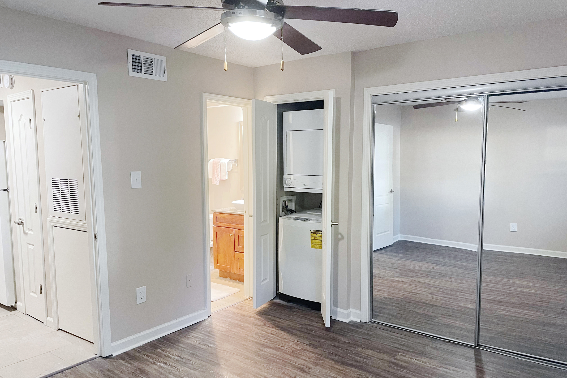 A light-filled room features a ceiling fan, a closet with sliding doors, and a laundry area visible through an open door. A bathroom is accessible nearby. The flooring is a wood-like laminate, and the walls are painted in a neutral color, creating a modern and spacious atmosphere.