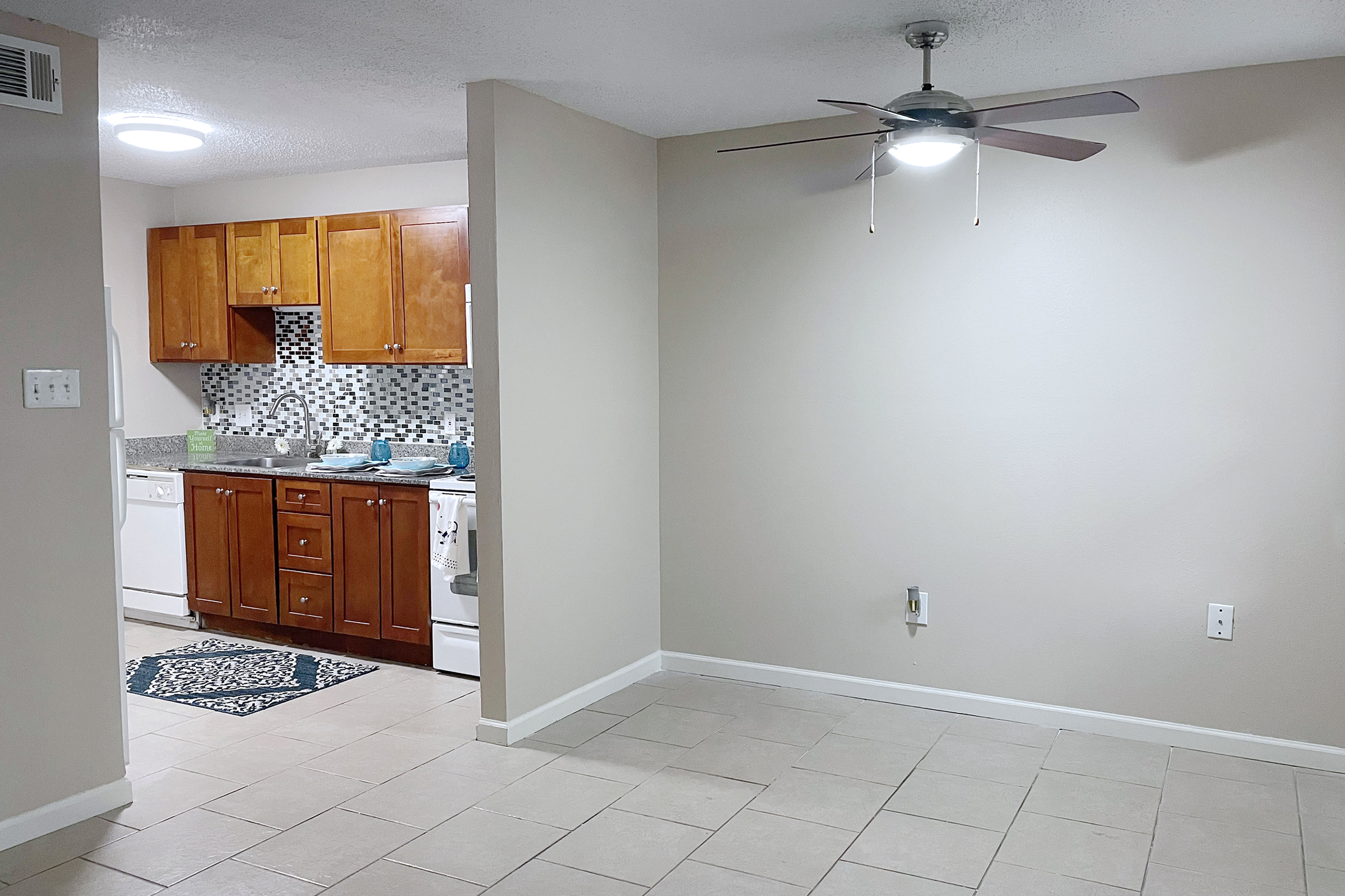 A brightly lit kitchen with wooden cabinets, a tiled backsplash, and a white dishwasher. Adjacent to the kitchen is an empty living space with light-colored walls and a ceiling fan. The floor is tiled, and a small area rug is visible in front of the kitchen.