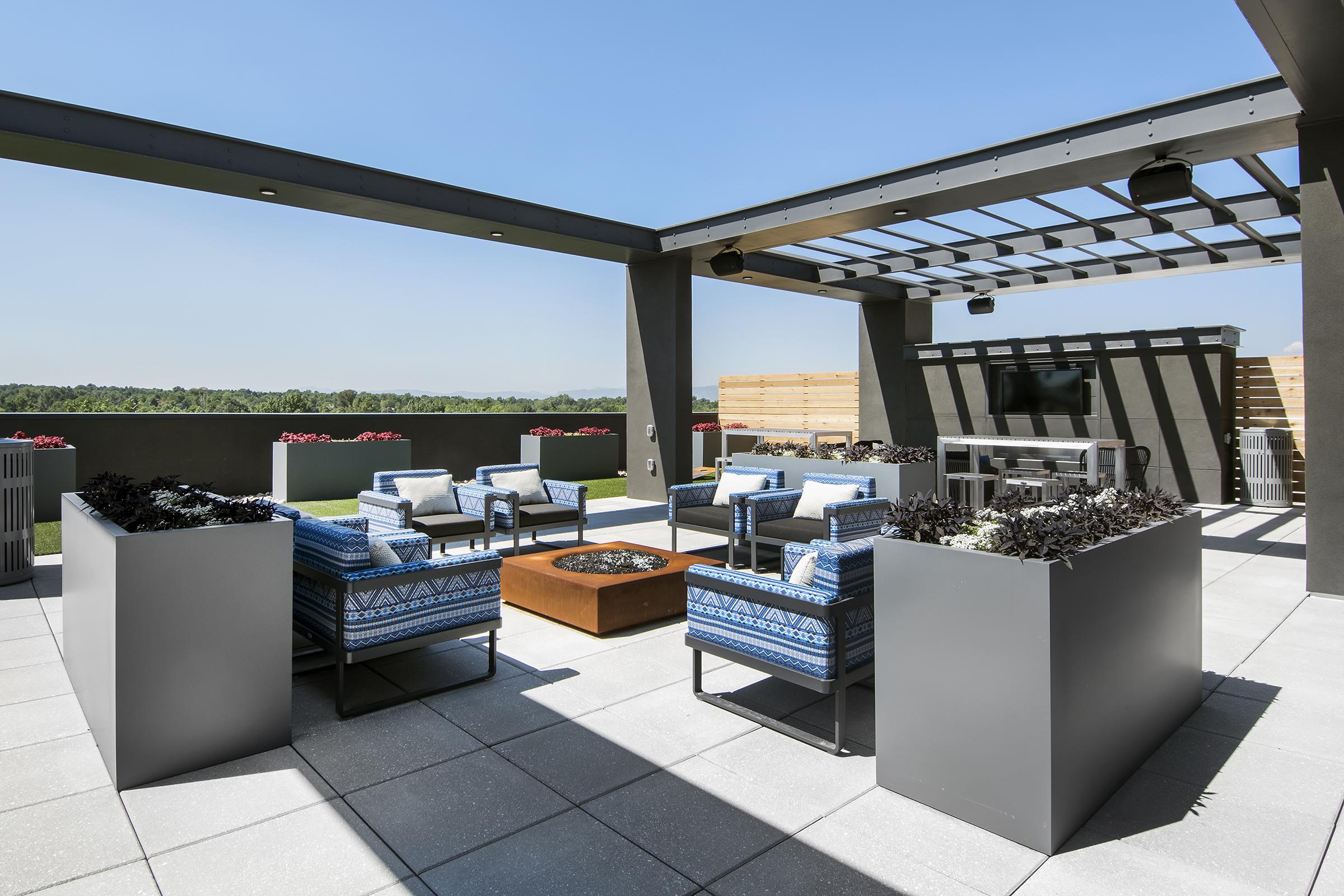 A modern rooftop terrace featuring comfortable blue-patterned seating arranged around a central fire pit. Planters with lush greenery decorate the space, and the area is bathed in sunlight under a clear blue sky. The backdrop includes a scenic view of green trees and distant landscape.