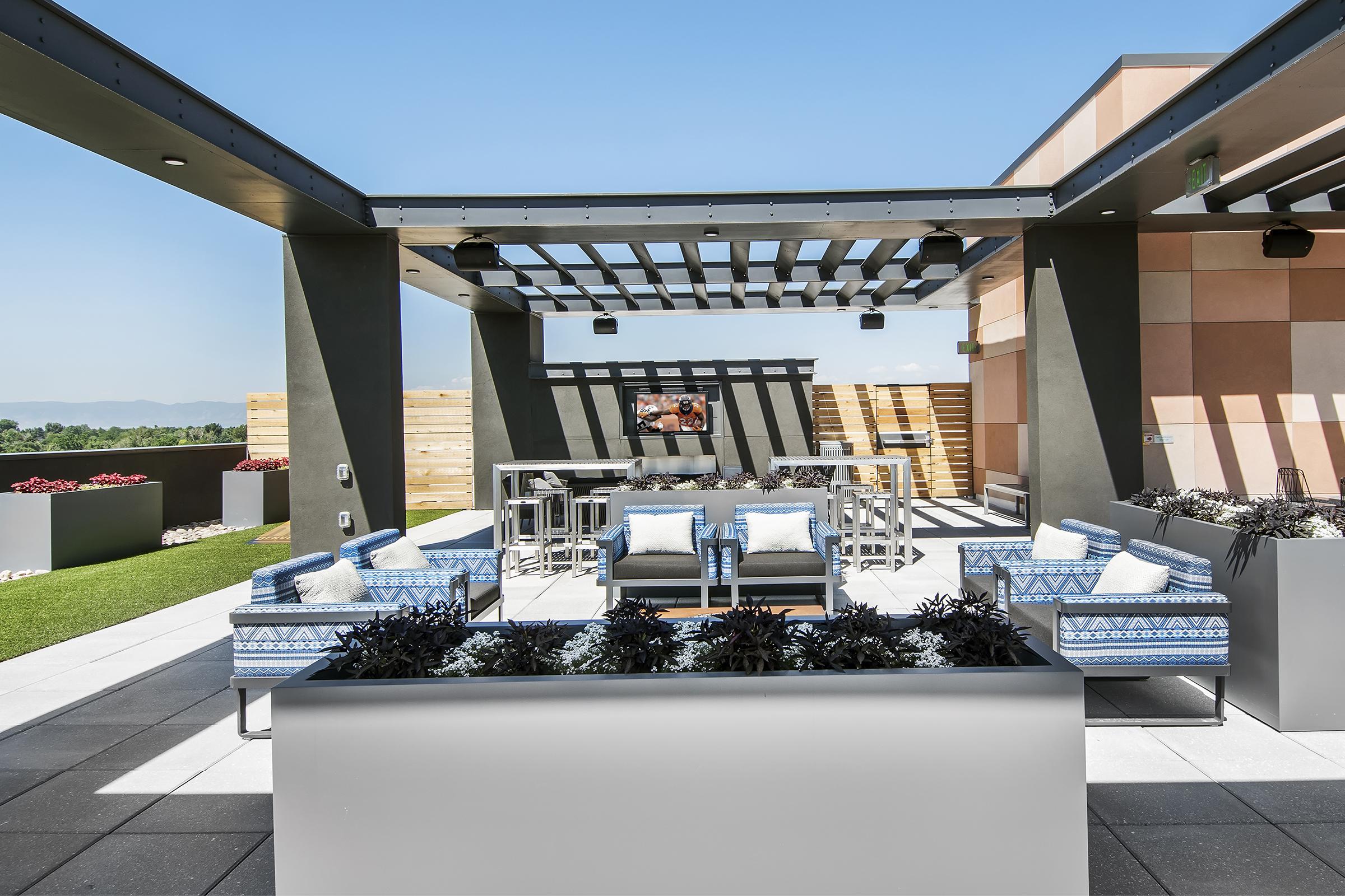 A modern rooftop lounge area featuring comfortable blue and white patterned seating, a bar table with high chairs, and decorative planters. In the background, a mounted TV is visible, surrounded by landscaped greenery and panoramic views of the landscape under a clear blue sky.
