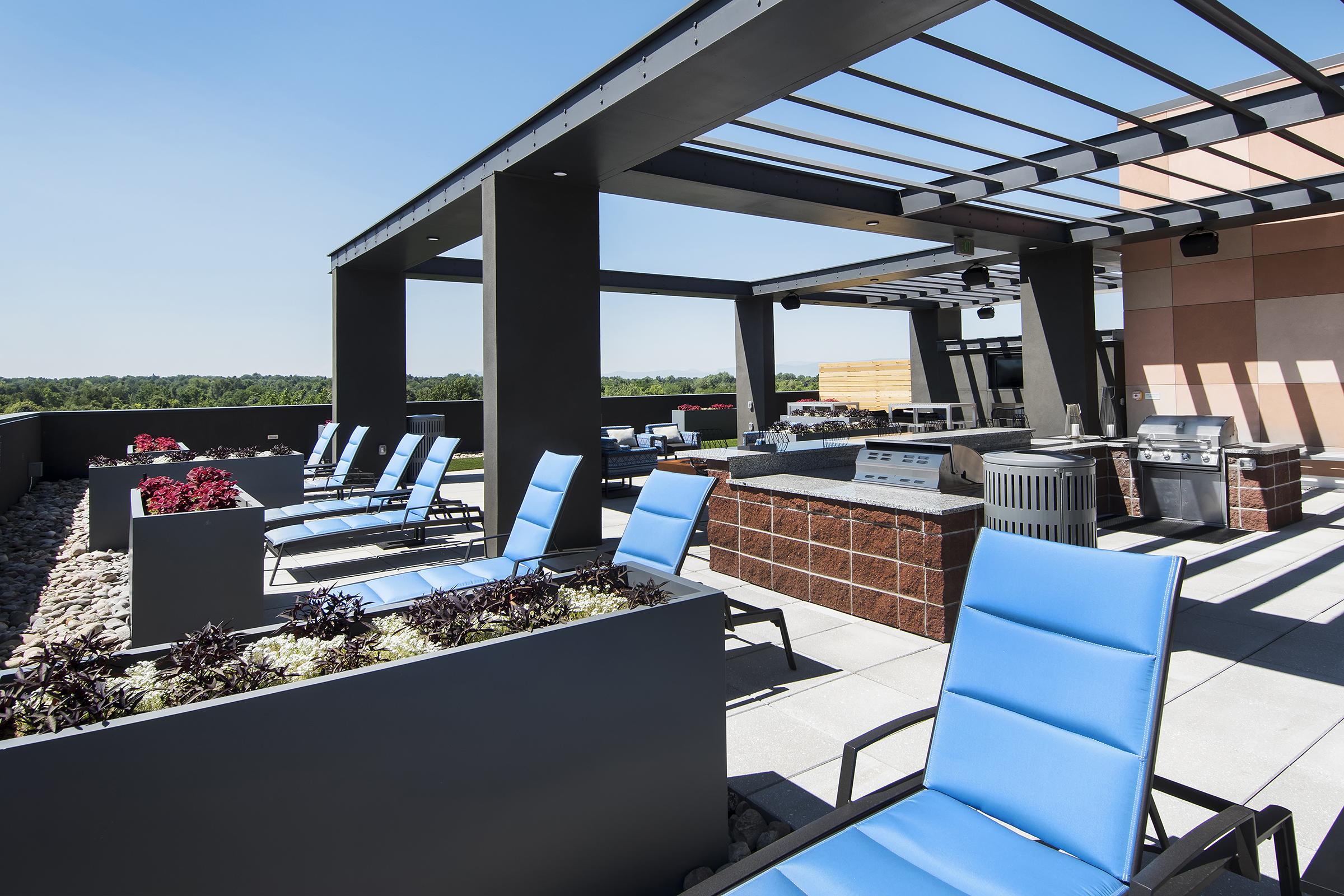Rooftop terrace featuring modern lounge chairs, open sky, and planters with greenery. A barbecue area is visible, along with outdoor cooking appliances, creating a stylish space for relaxation and socializing. The surrounding landscape is visible in the distance.