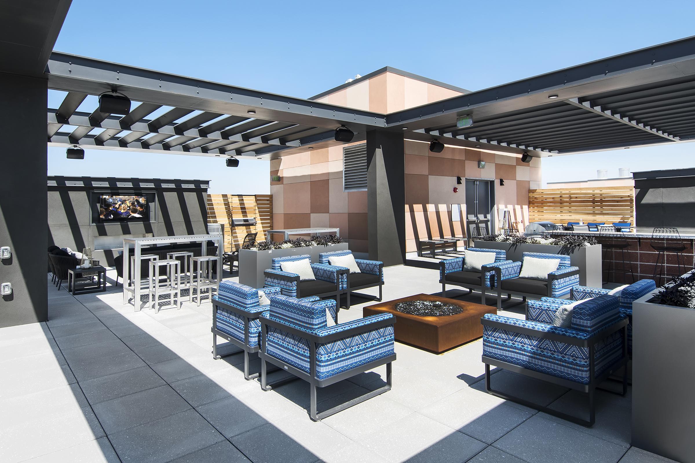 A modern rooftop lounge area featuring stylish blue patterned furniture, a fire pit, and a large TV screen. The space is open-air, with metal beams overhead and bright sunlight, creating a relaxing atmosphere. There are also tables and seating arrangements for social gatherings.
