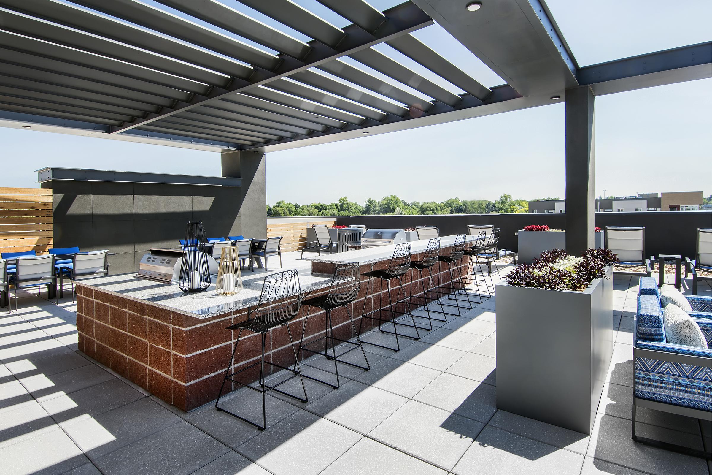Rooftop outdoor lounge area featuring a modern bar with seating, surrounded by planters and open space. The area has a slatted overhead structure allowing sunlight to filter through, creating a stylish and inviting atmosphere for relaxation and socializing. Clear skies in the background enhance the setting.