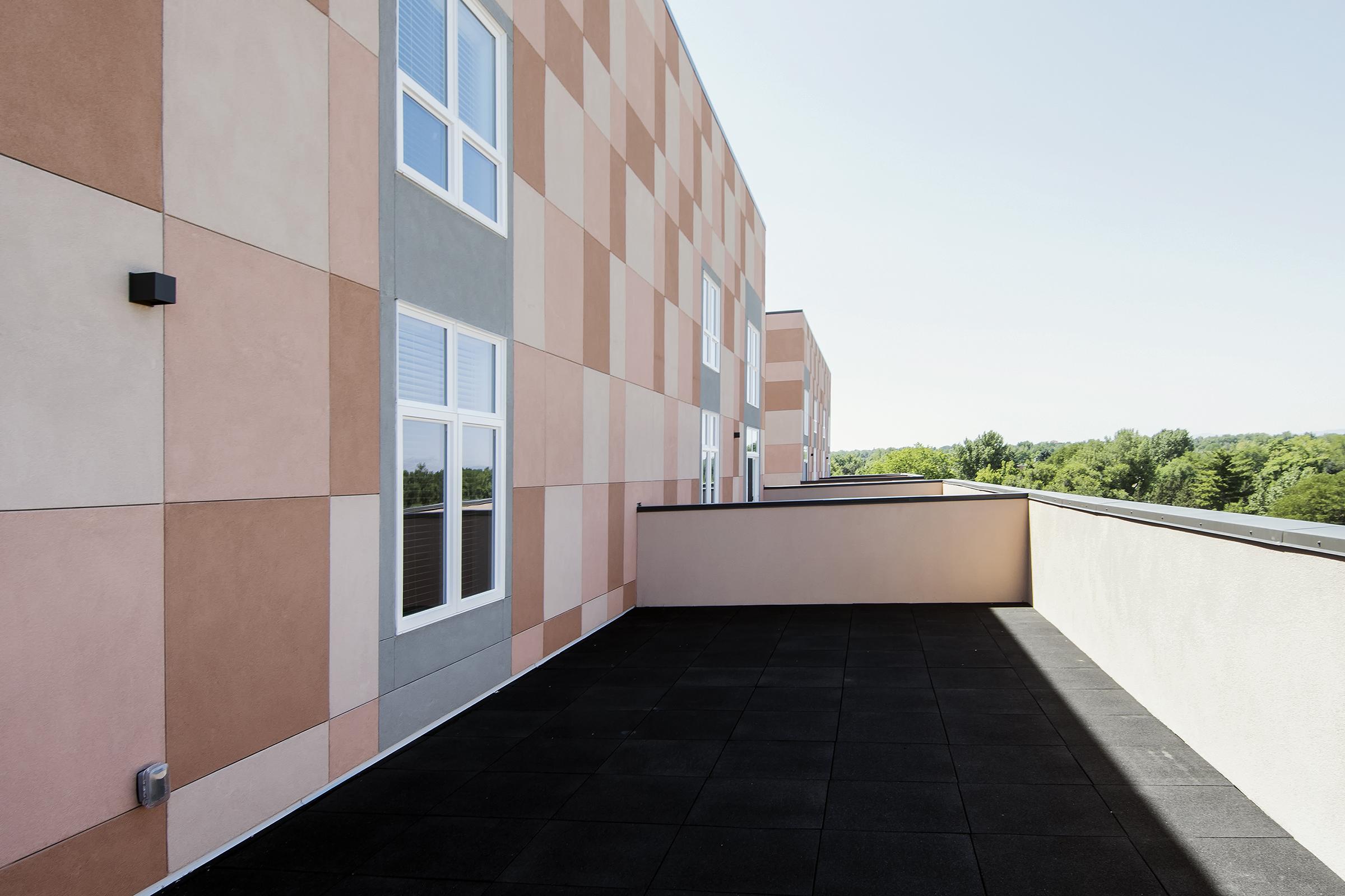 A modern balcony with a flat black surface, surrounded by a building featuring a pattern of pastel-colored panels in pink, gray, and beige. The landscape in the background includes trees under a clear blue sky.
