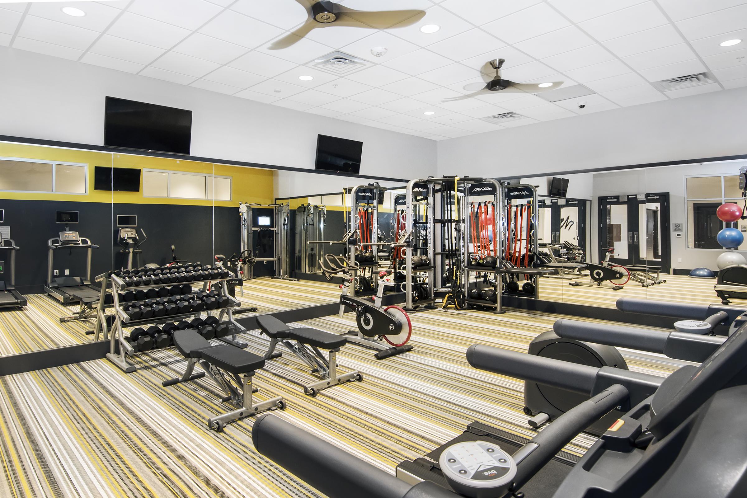A modern gym interior featuring a variety of workout equipment, including weight benches, dumbbells, and resistance machines. The space is brightly lit with overhead lights and large mirrors, and has striped flooring. Multiple television screens are mounted on the walls, enhancing the workout environment.