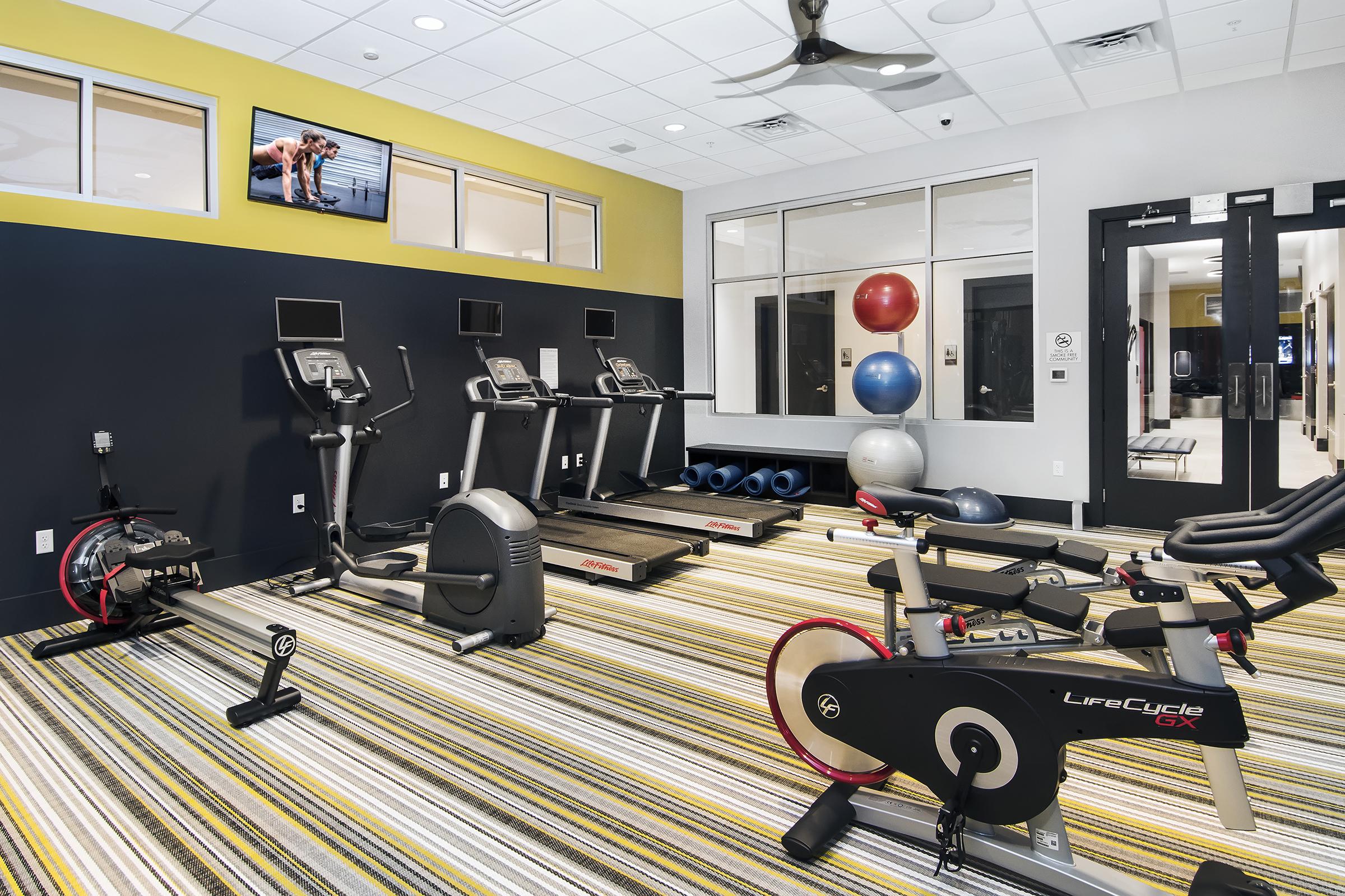A modern gym interior featuring several exercise machines, including treadmills, an elliptical, and a stationary bike. There are colorful exercise balls and wall-mounted screens displaying workout videos. The room is well-lit with a striped carpet and a ceiling fan.