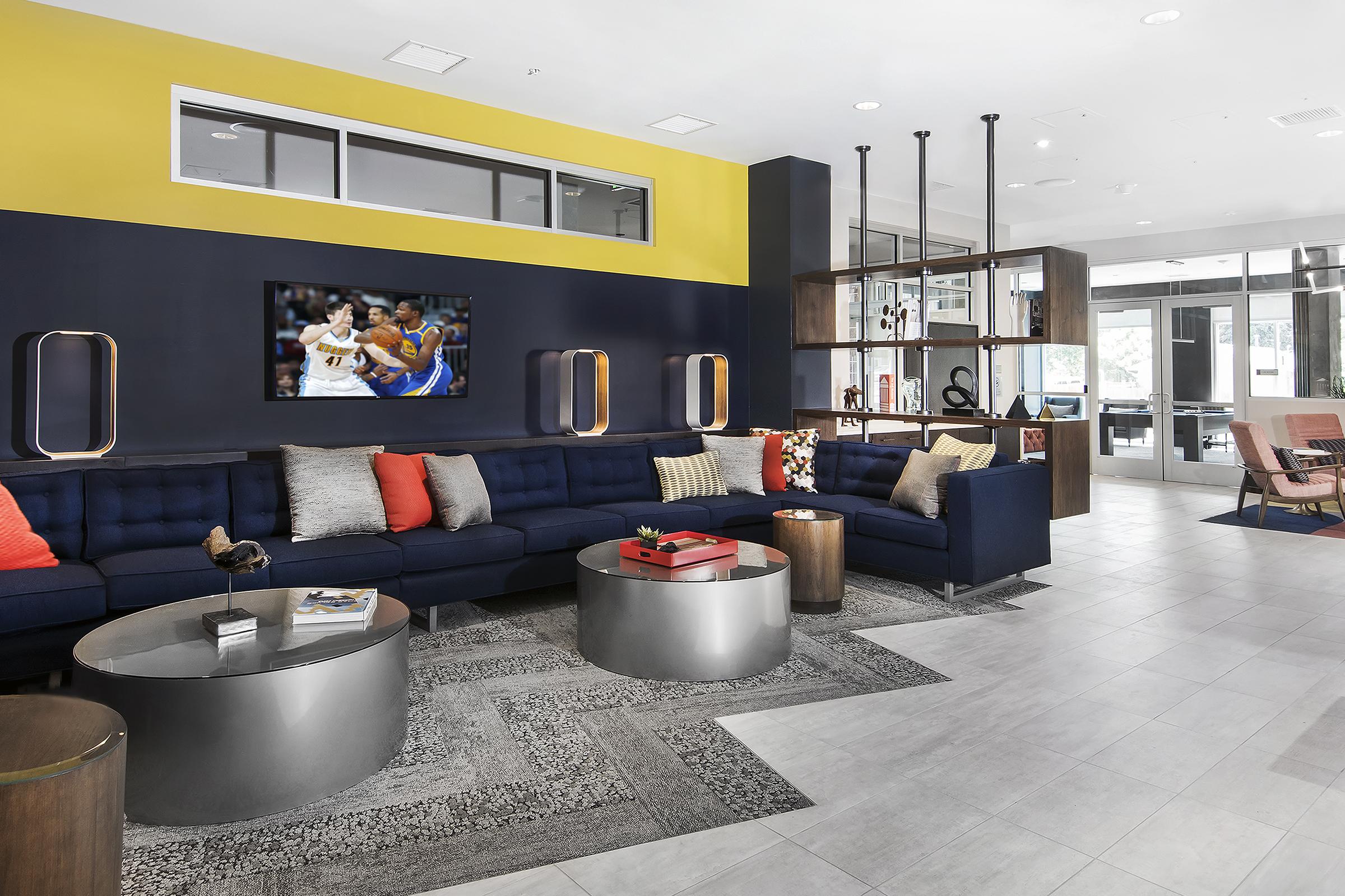 A modern lounge area featuring a navy blue and yellow color scheme. There are two large sofas with colorful cushions, a round coffee table, and a wall-mounted TV displaying a sports game. The space includes stylish decor elements and large windows letting in natural light.