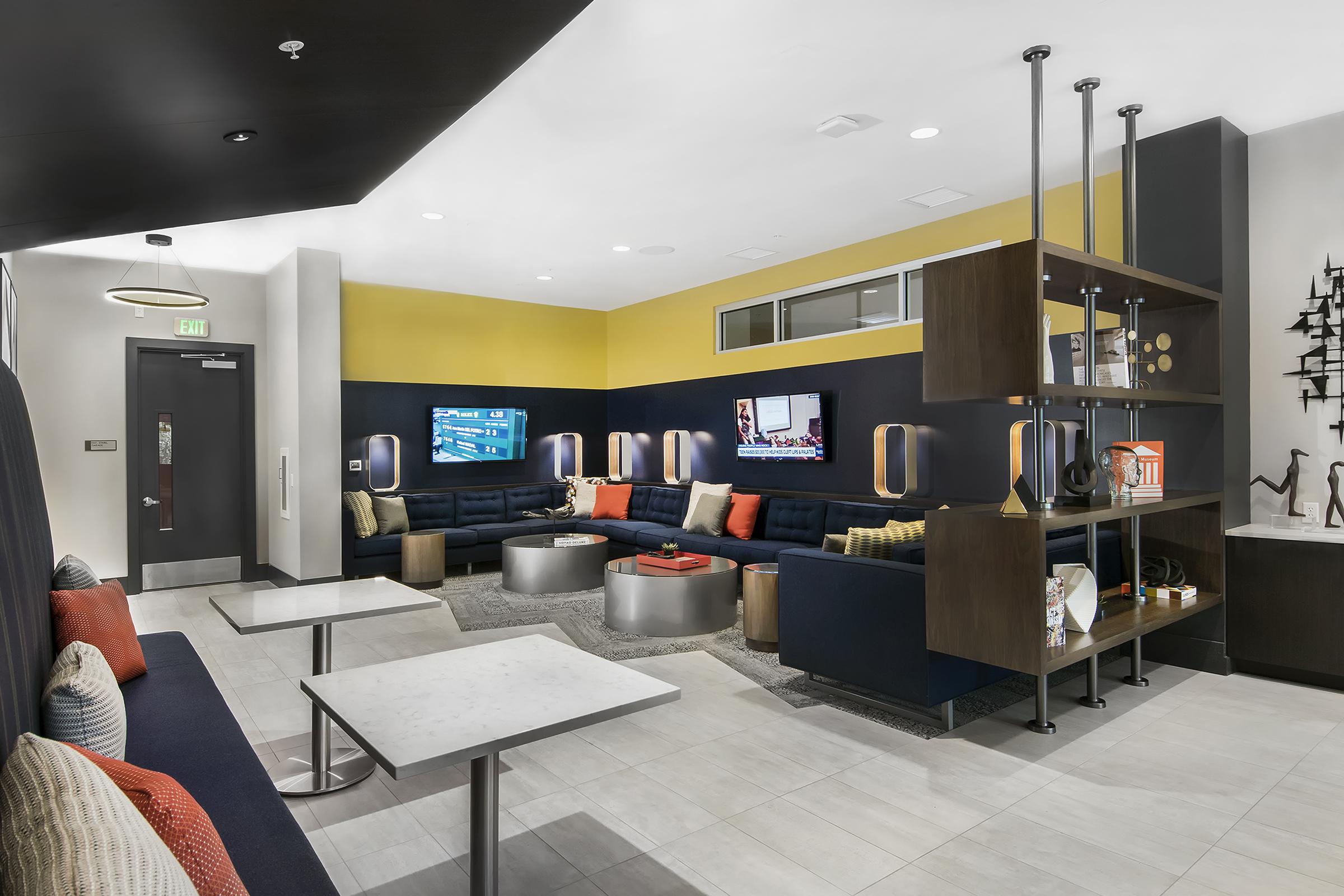A modern lounge area featuring comfortable seating with colorful cushions, a stylish coffee table, and multiple flat-screen TVs mounted on the walls. The space is designed with a contemporary aesthetic, incorporating yellow and dark blue accents, and includes decorative shelves with various items.