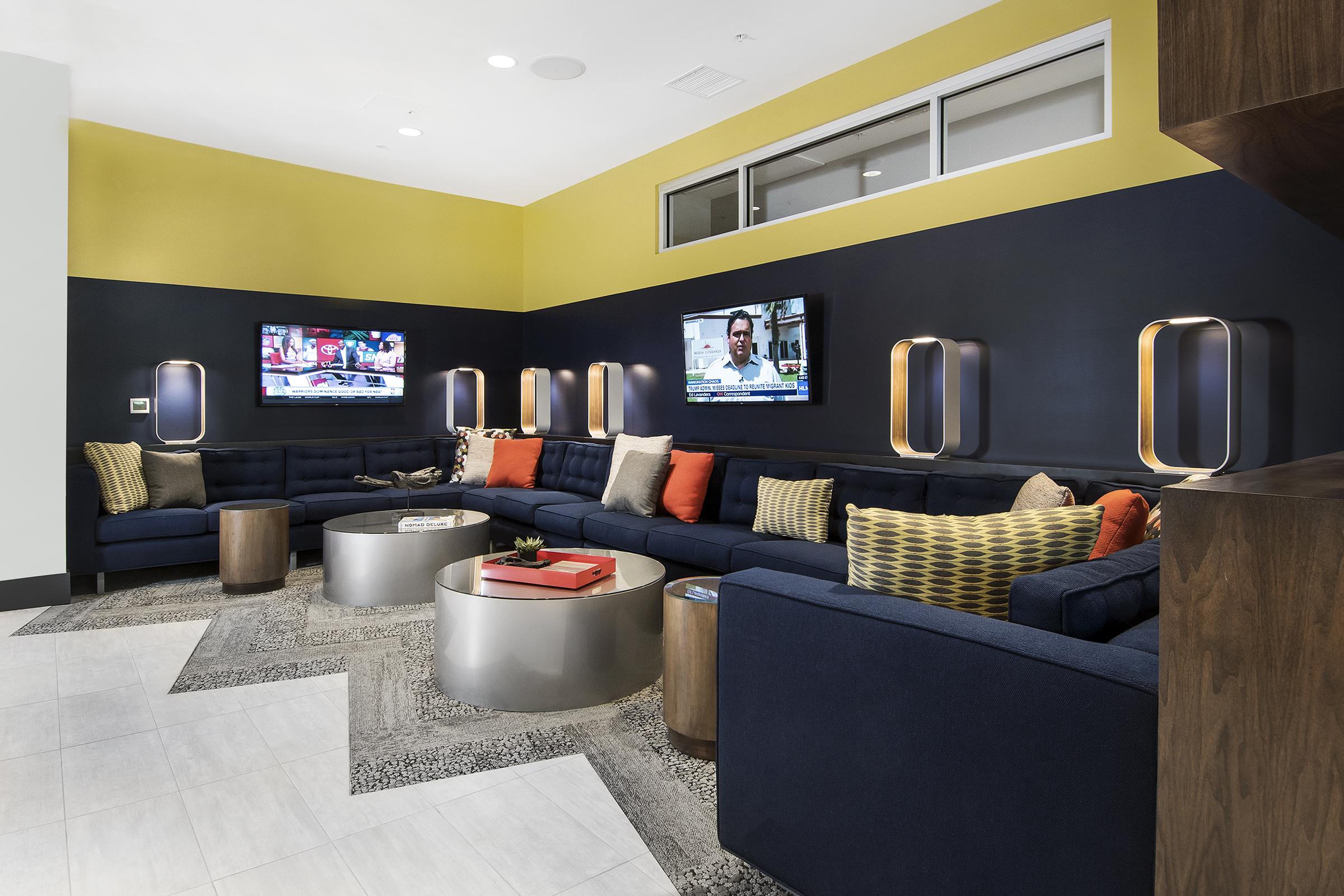 A modern lounge area featuring a large navy blue sectional sofa adorned with colorful throw pillows. Two flat-screen TVs mounted on the wall show different programs. The space includes round coffee tables made of wood and glass, with decorative elements, and walls painted in a combination of yellow and dark blue.