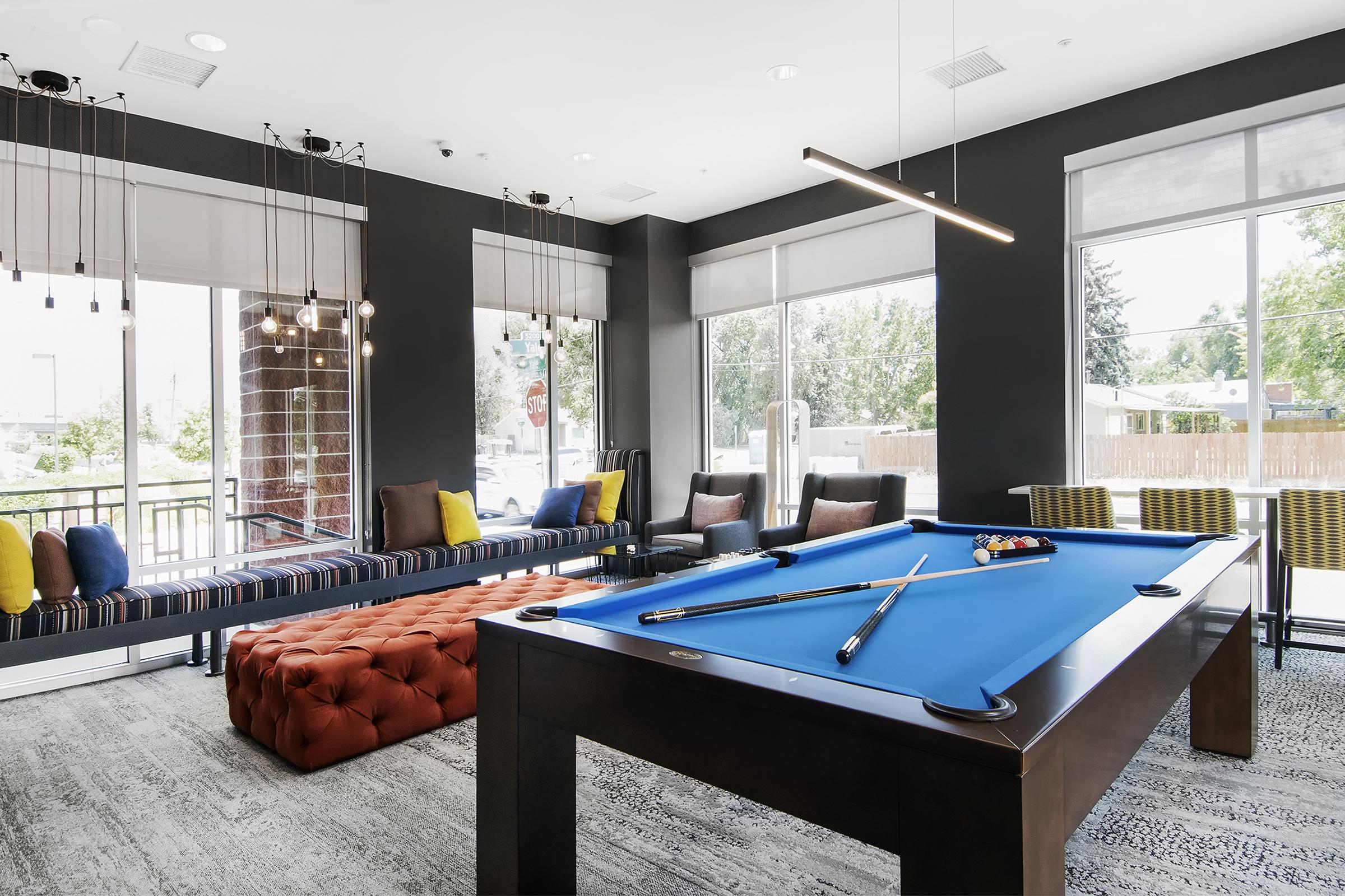 A modern leisure area featuring a billiards table with a blue felt surface, surrounded by stylish seating. Soft natural light filters through large windows, illuminating the space, which includes a cozy seating arrangement with colorful cushions and a large ottoman.