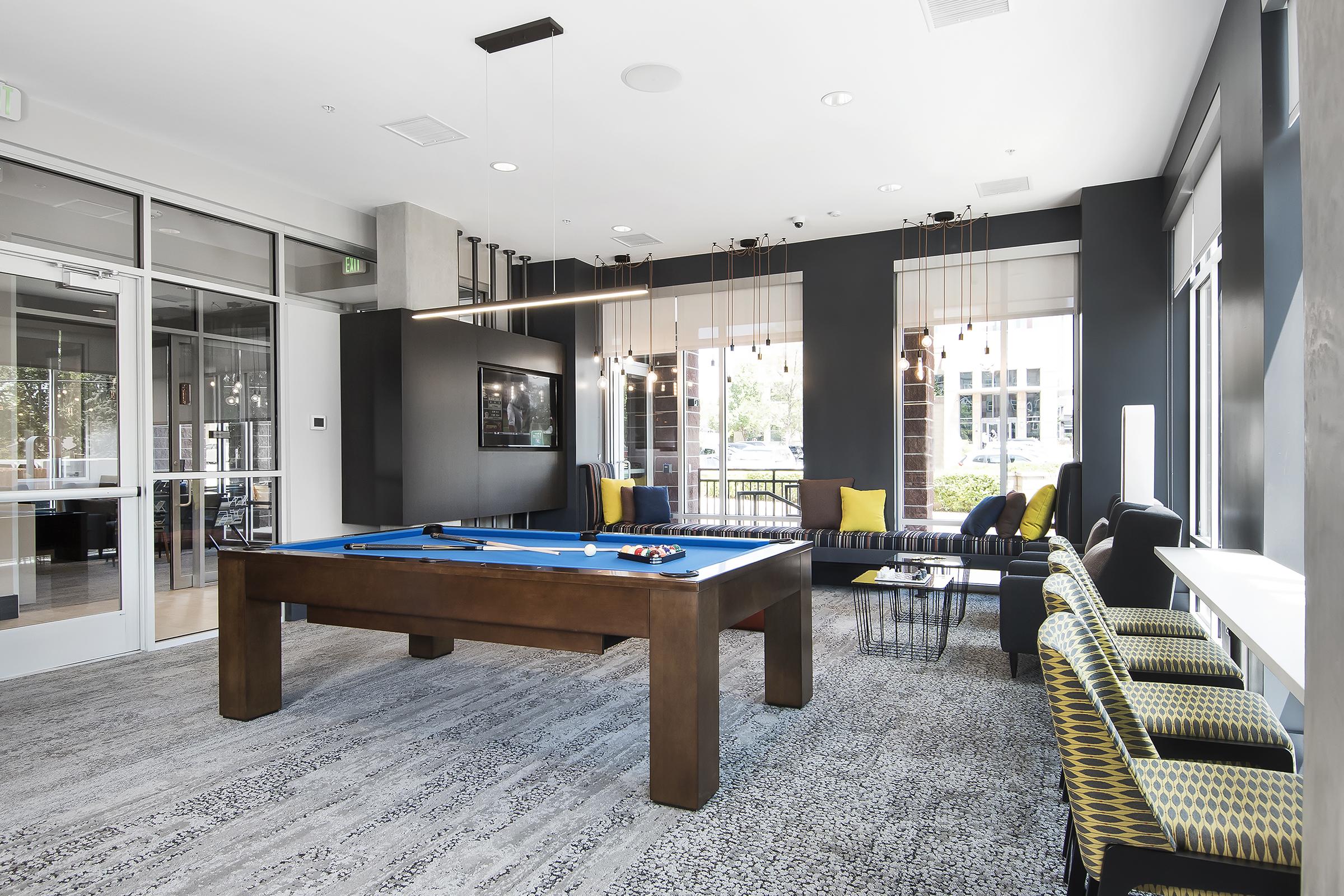 A modern game room featuring a billiards table with a blue felt surface, comfortable seating areas with yellow cushions, and large windows allowing natural light. The decor includes contemporary lighting fixtures and a large TV mounted on the wall, creating a welcoming and stylish atmosphere for socializing and relaxation.
