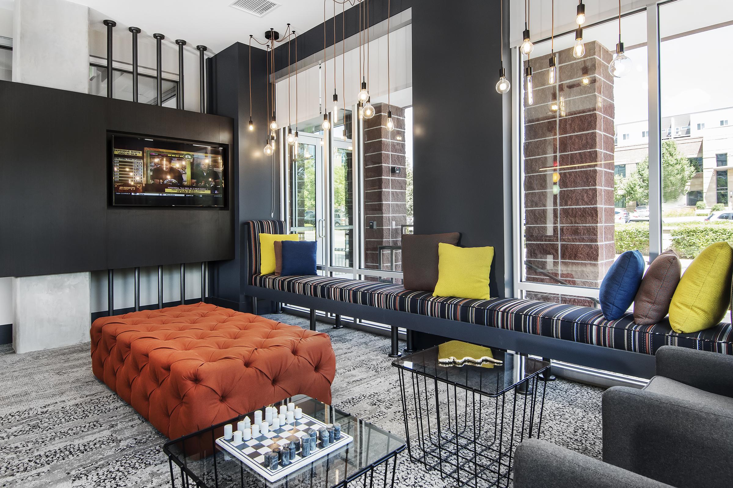 A modern lounge area featuring a large orange tufted ottoman, striped bench seating with colorful cushions, and sleek black metal coffee tables. The space is illuminated by hanging light fixtures and includes a TV mounted on the wall, with large windows allowing natural light to enter.