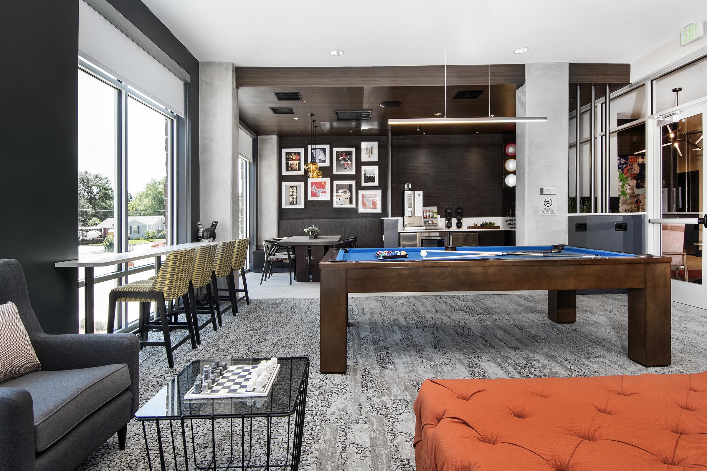 A modern lounge area featuring a cozy seating arrangement with a gray sofa and orange ottoman. There's a billiards table, a chess table, and large windows allowing natural light. The walls are adorned with artwork, and there's a bar-style seating area in the background.