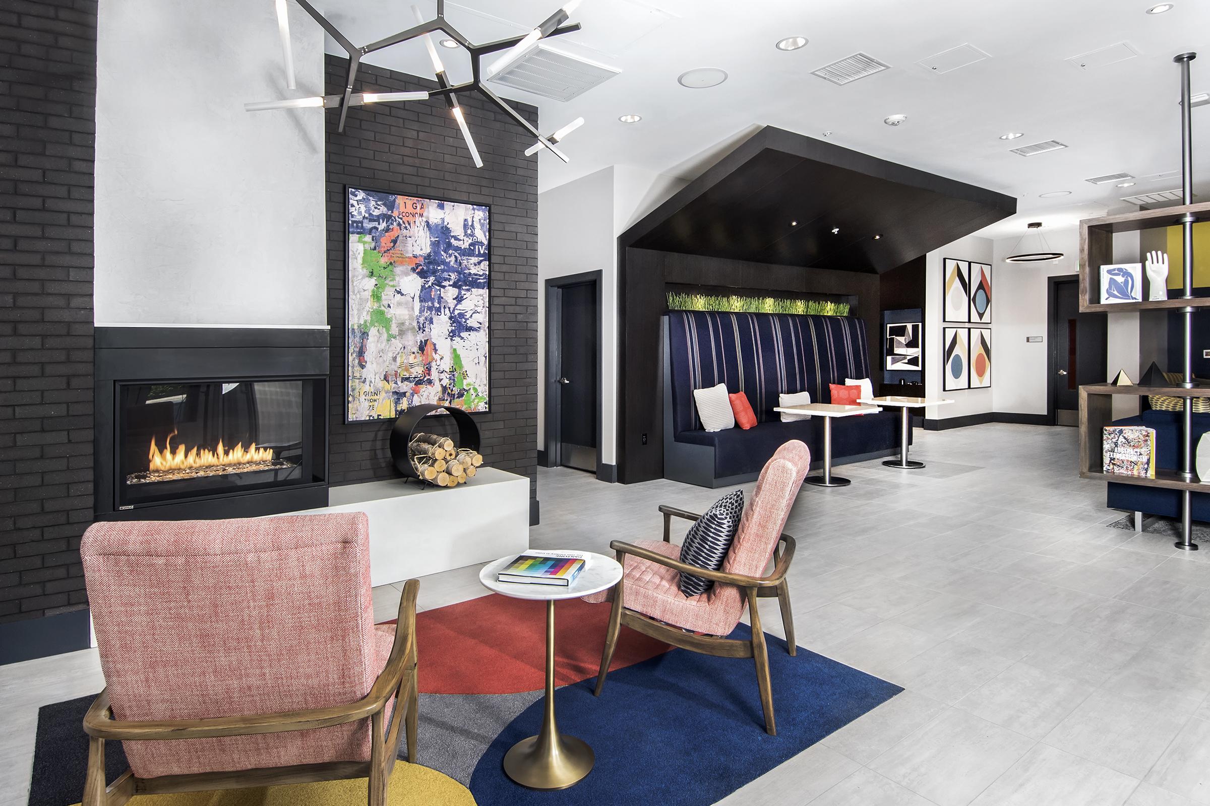 A modern lobby featuring a cozy fireplace, two colorful armchairs, and contemporary artwork on the walls. There's a mix of seating areas, including a booth with navy blue upholstery, and a stylish coffee table with magazines. The space is well-lit with a contemporary chandelier and has a welcoming atmosphere.