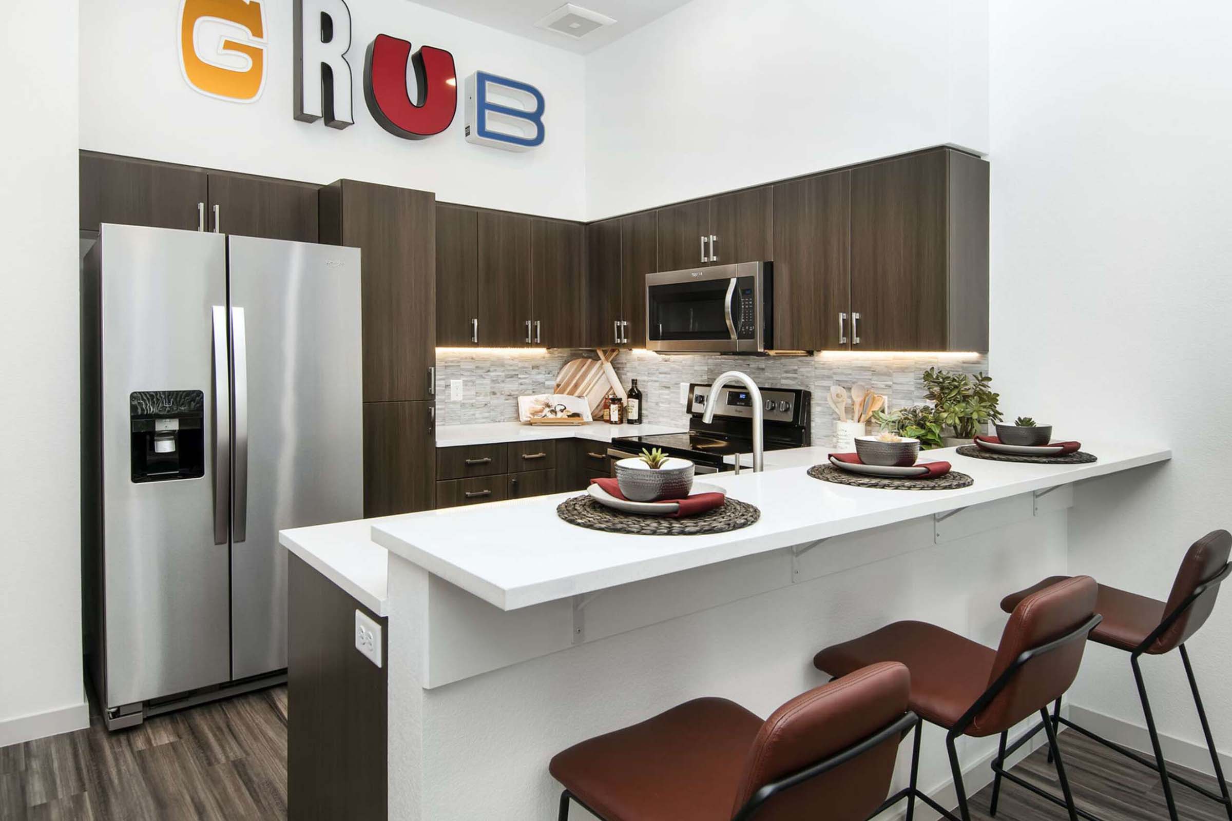 Modern kitchen featuring dark wood cabinets, stainless steel appliances, and a white countertop. A large silver refrigerator is visible, along with a microwave and stove. The kitchen has decorative plants and table settings on the countertop, with red and blue letters spelling "GRUB" on the wall above.