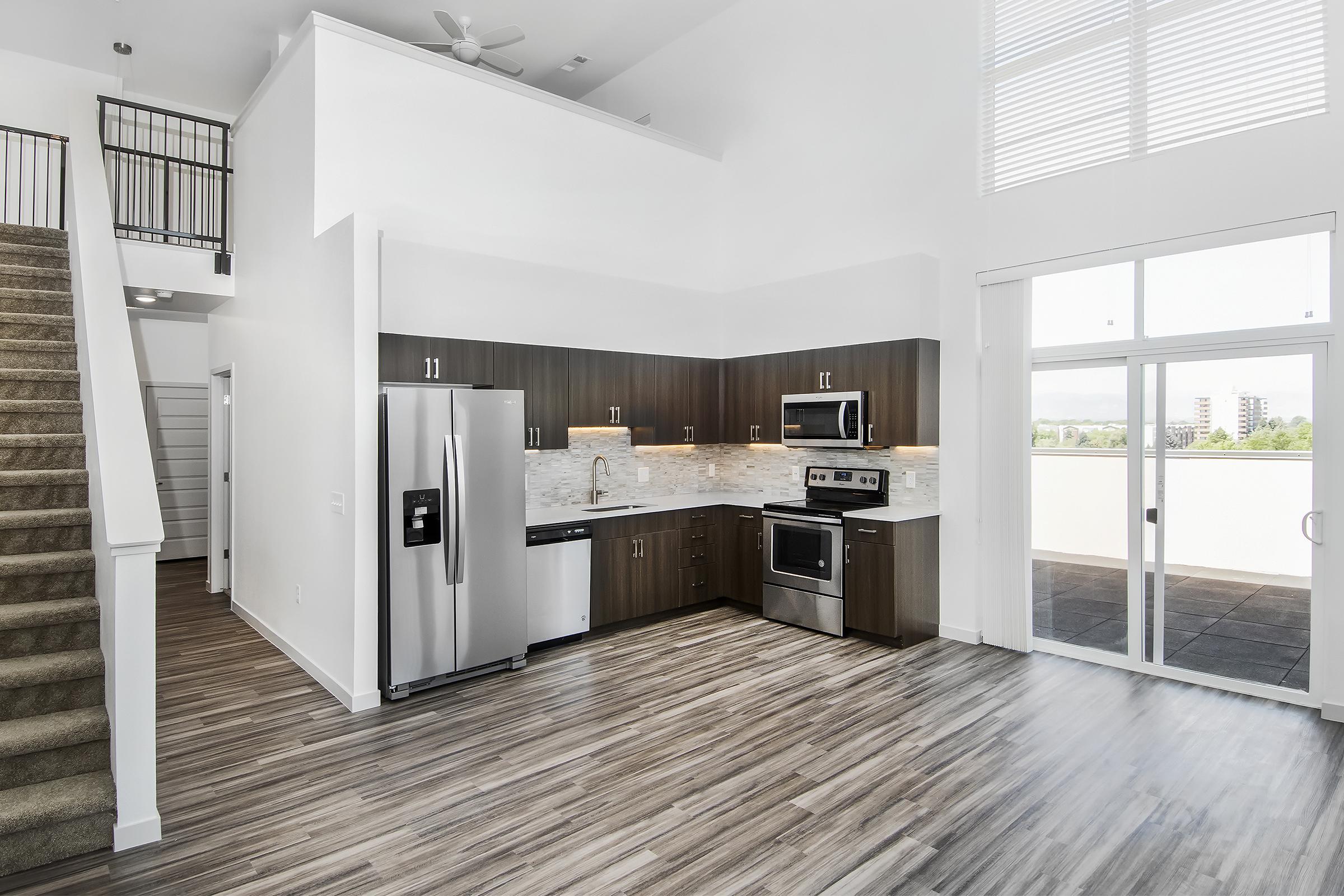 Modern kitchen and living area featuring stainless steel appliances, dark wood cabinetry, and marble countertops. Large windows provide natural light, with a staircase leading to an upper level. Floor is covered in wood-like laminate, creating a warm and inviting atmosphere.