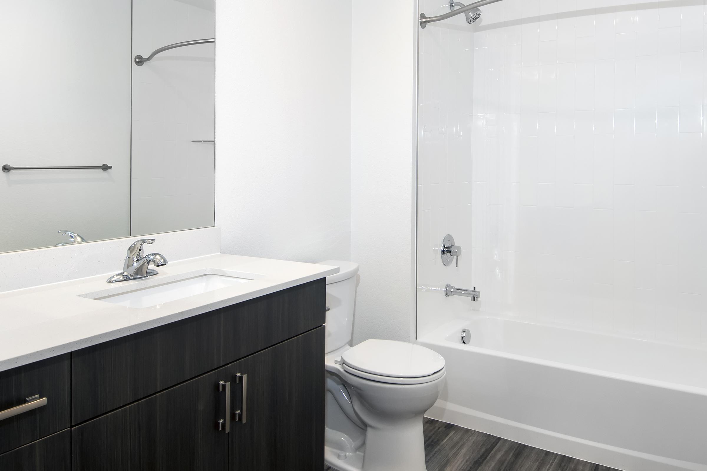 A modern bathroom featuring a white bathtub and shower combination, a sleek sink with a dark vanity, and a toilet. The walls are painted white, and there is a large mirror above the sink. The floor has a dark wood-like finish, creating a clean and contemporary aesthetic.