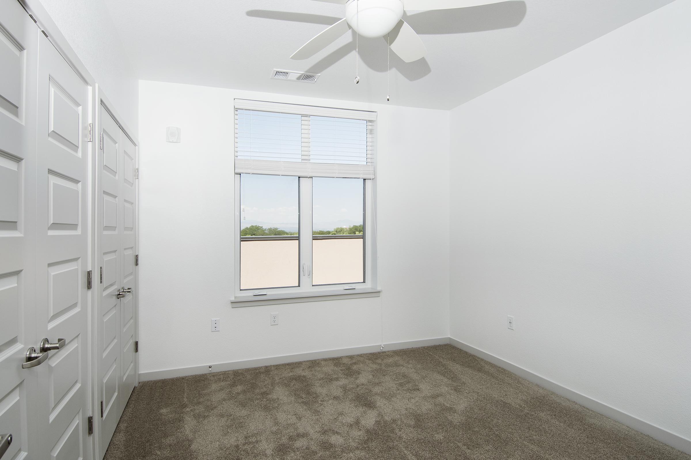 A well-lit, empty room with beige carpet, two doors, and a window featuring blinds. A ceiling fan is visible overhead. The walls are painted white, contributing to a spacious and minimalist atmosphere.