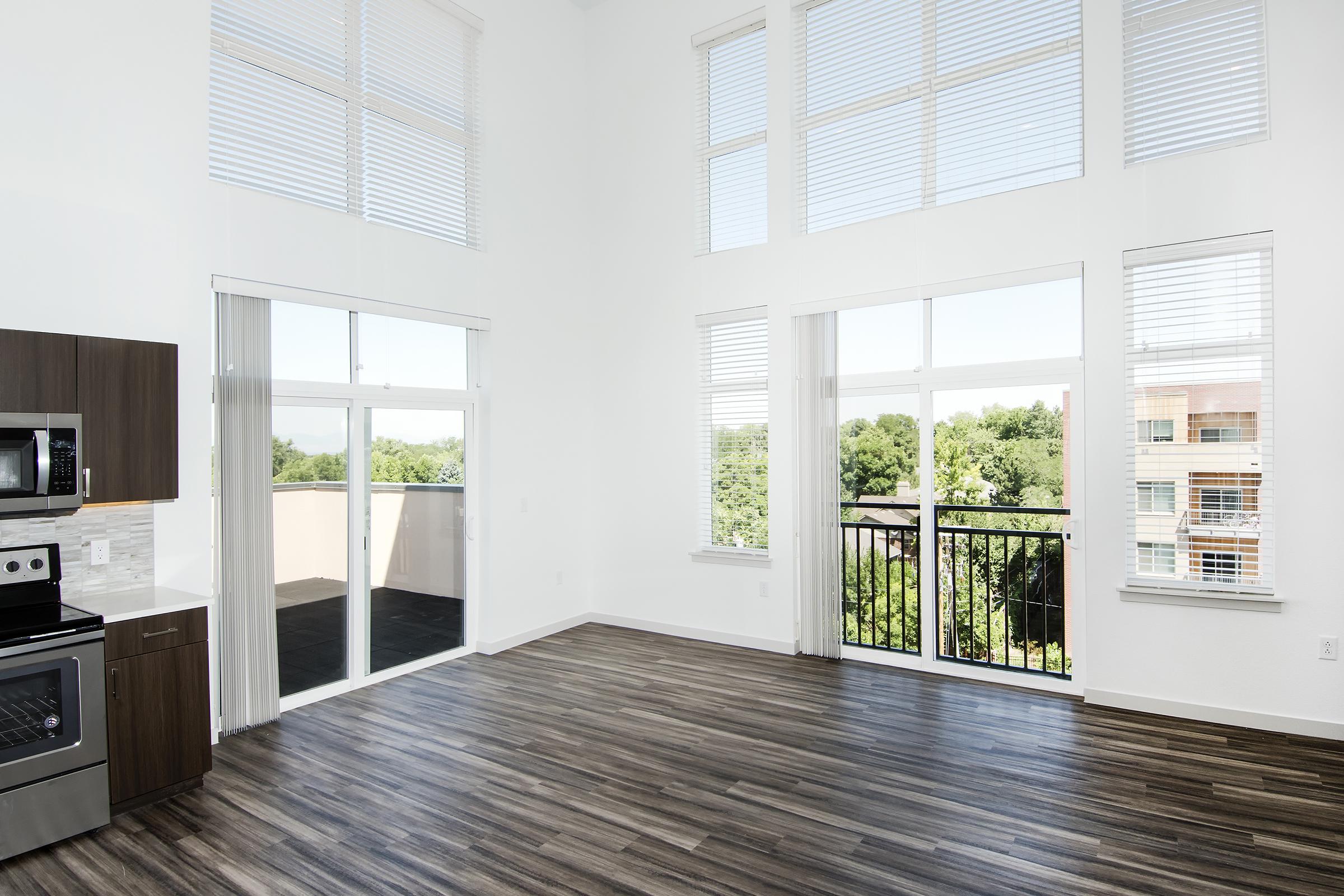 Bright and spacious interior of a modern apartment featuring large windows with natural light, wood-like flooring, and an open kitchen area with stainless steel appliances. A balcony with railing offers a view of greenery outside the apartment.