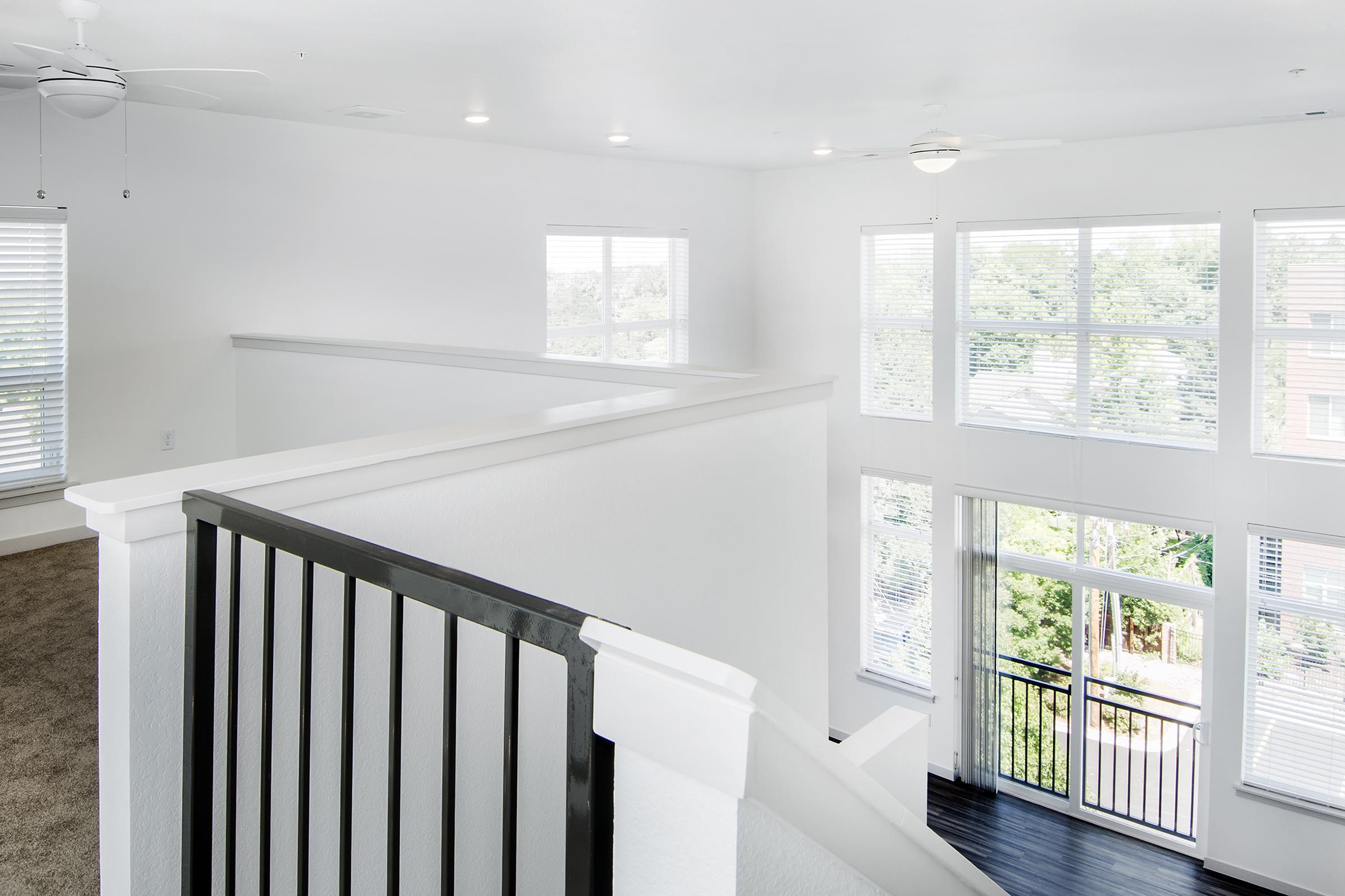 Bright, modern interior of a home featuring a spacious living area with large windows allowing natural light. The room has white walls, a ceiling fan, and a staircase with a black railing. A carpeted area is visible alongside the wooden flooring, creating a cozy yet elegant atmosphere.